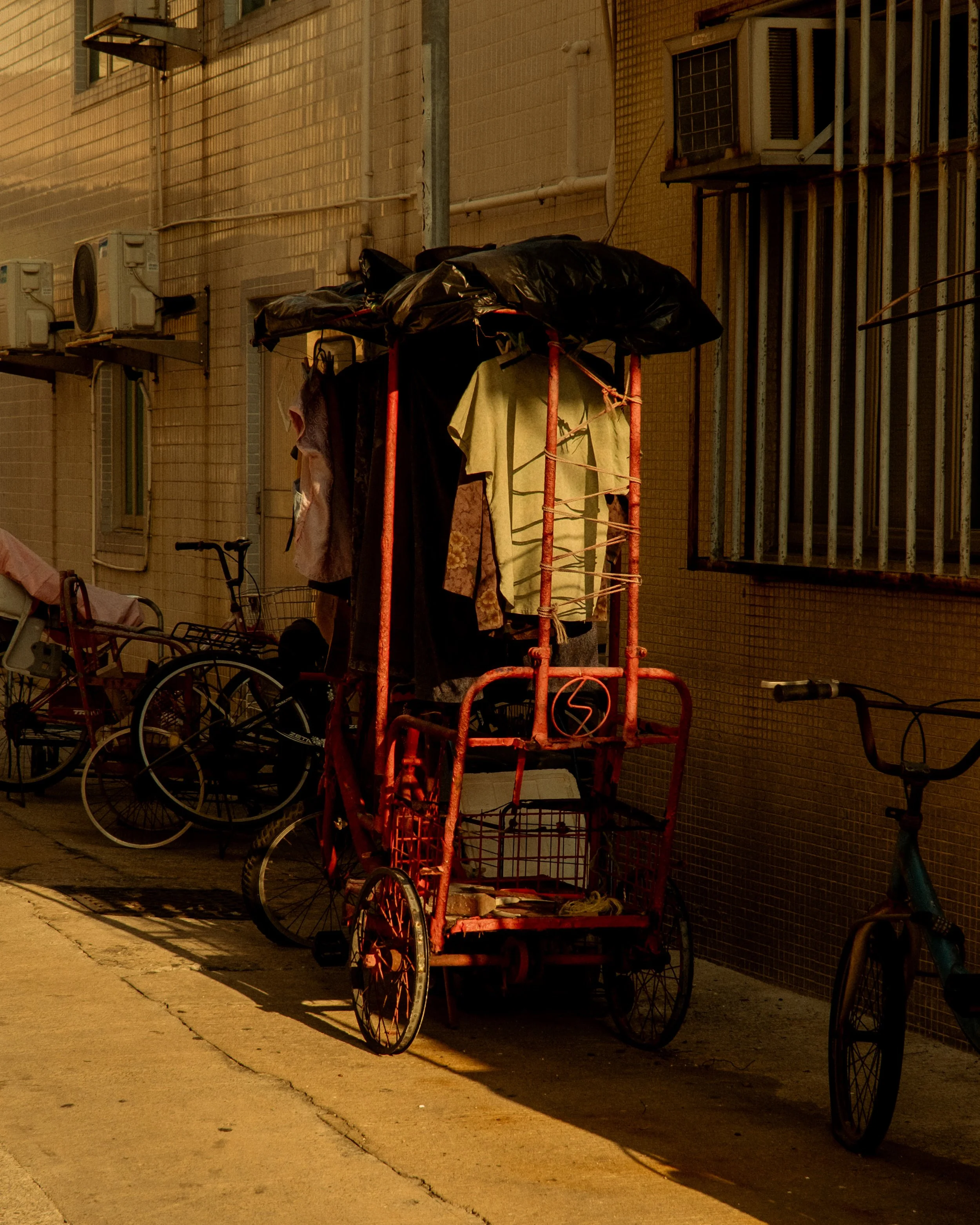A red pedicab parked on the sidewalk near a beige building with barred windows and air conditioning units, with clothes hanging on a line attached to the pedicab.