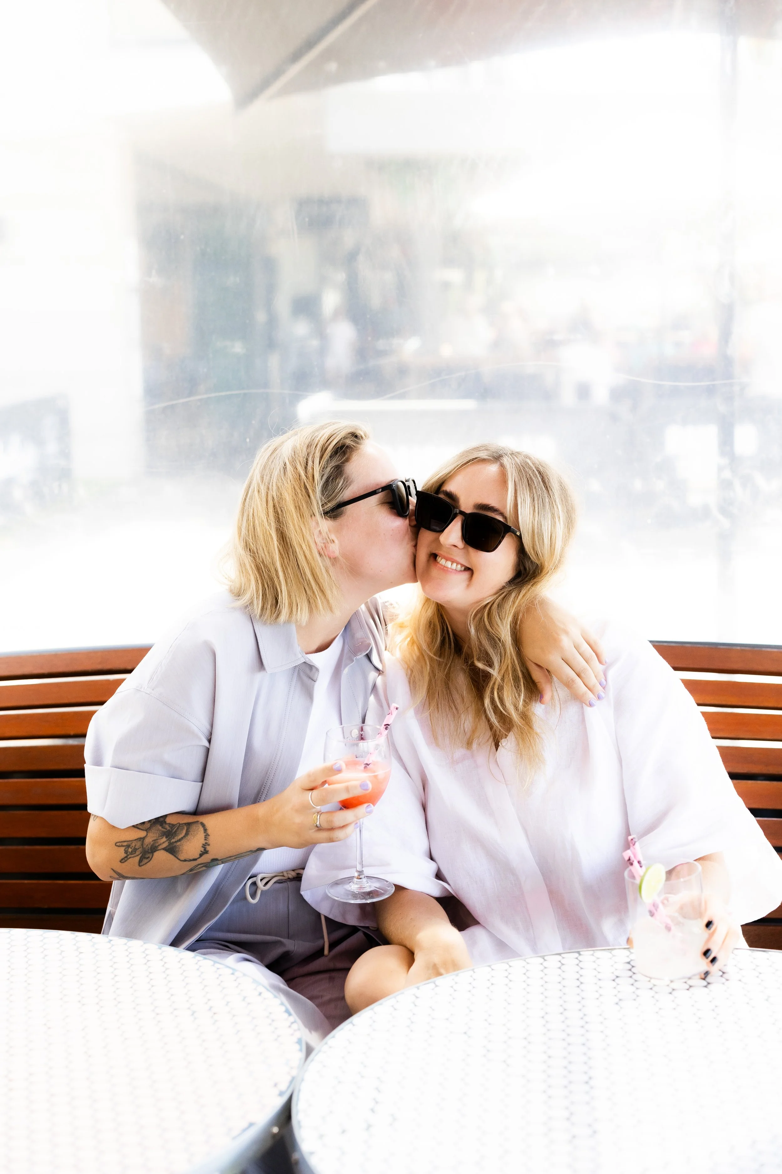Two women wearing sunglasses sharing a kiss on the cheek at an outdoor cafe, holding drinks, sitting on a wooden bench with metal tables in front and a glass window behind them.