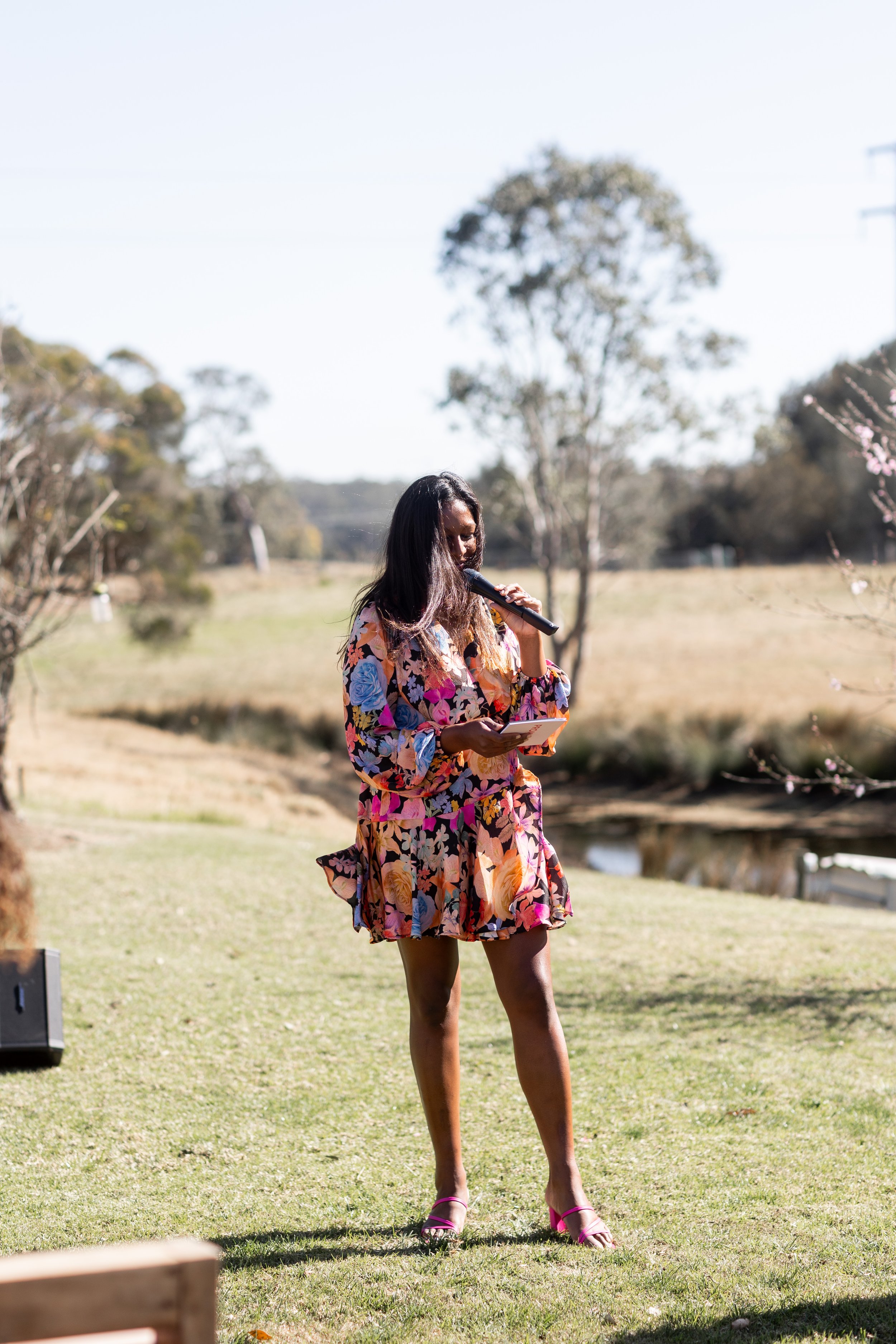 A woman in a colorful floral dress stands outdoors on grass, holding a microphone and a piece of paper, possibly speaking or reading to an audience at an event or gathering.