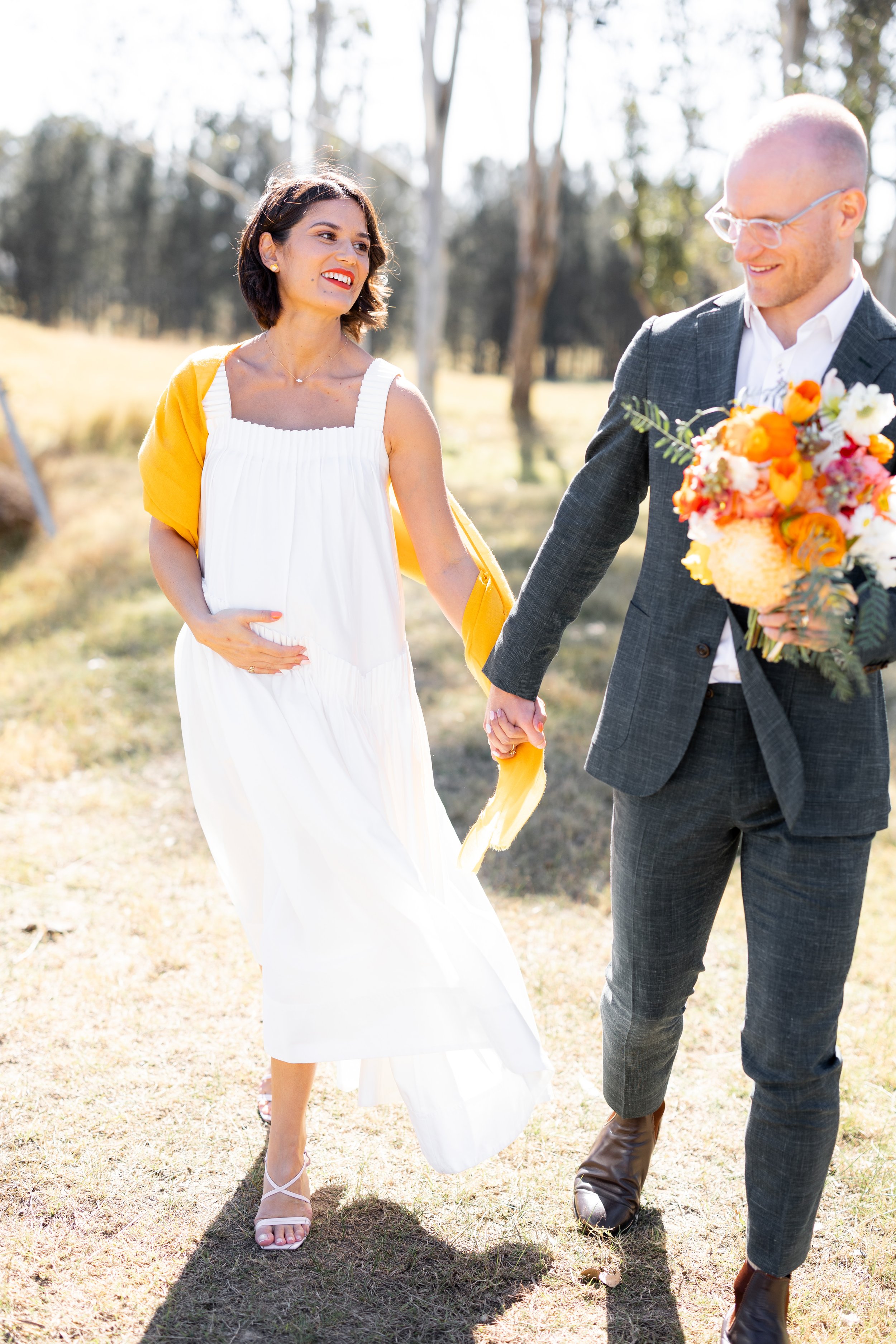 A couple holding hands outdoors during a wedding. The woman wears a white dress with a yellow shawl, and the man wears a grey suit and glasses. The man holds a bouquet of orange, pink, and white flowers.