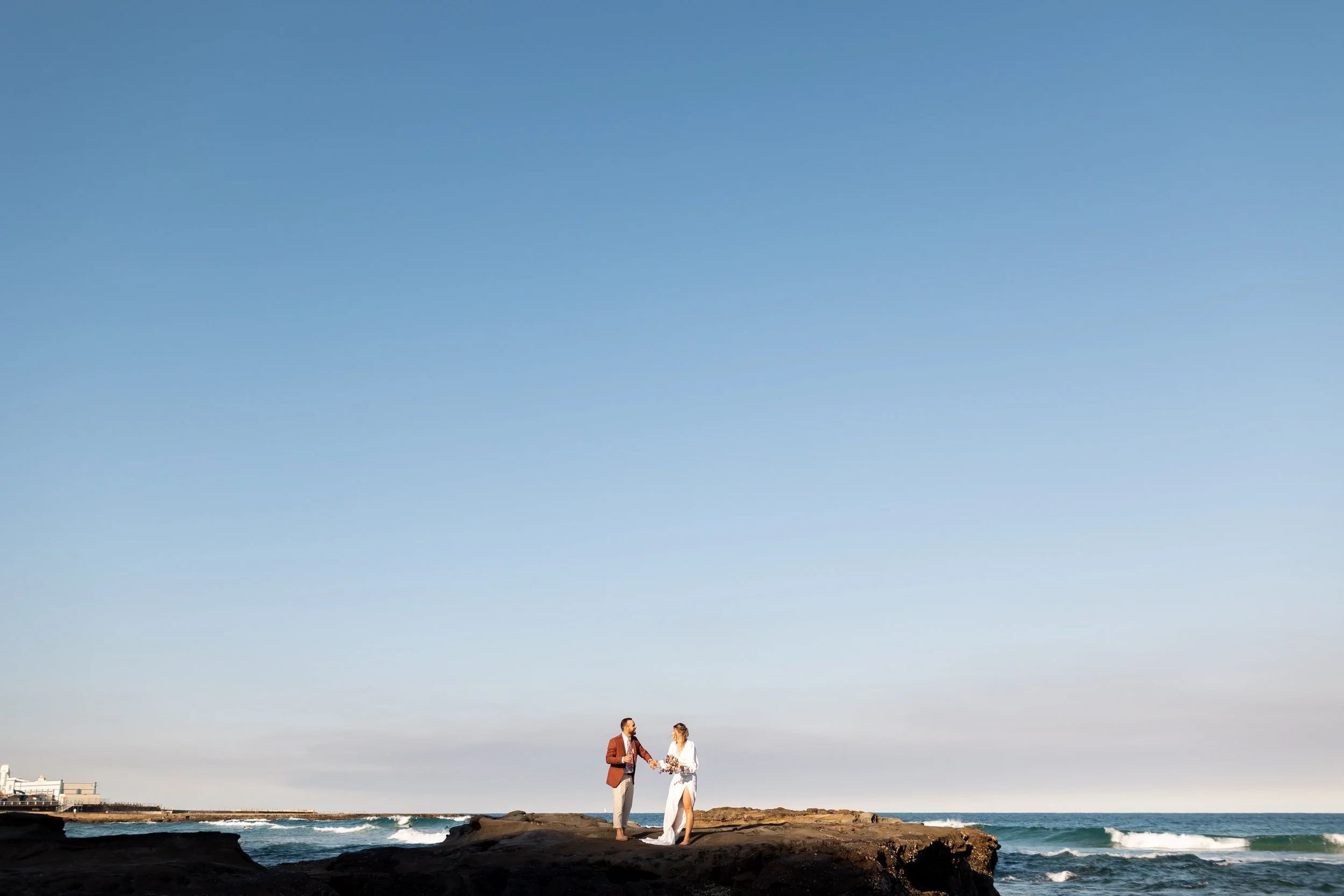 A couple dressed in formal attire standing on a rocky point by the ocean, exchanging vows during a wedding ceremony, with clear blue sky and waves crashing in the background.