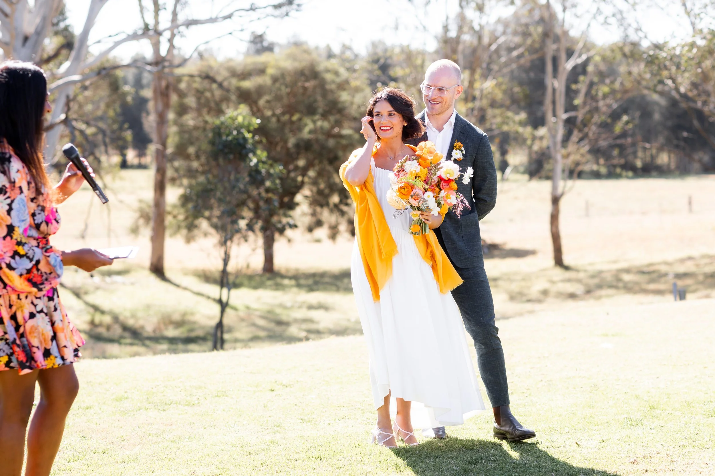 A couple stands outside during a wedding ceremony, with a woman holding a bouquet of vibrant flowers, smiling and looking at the officiant.