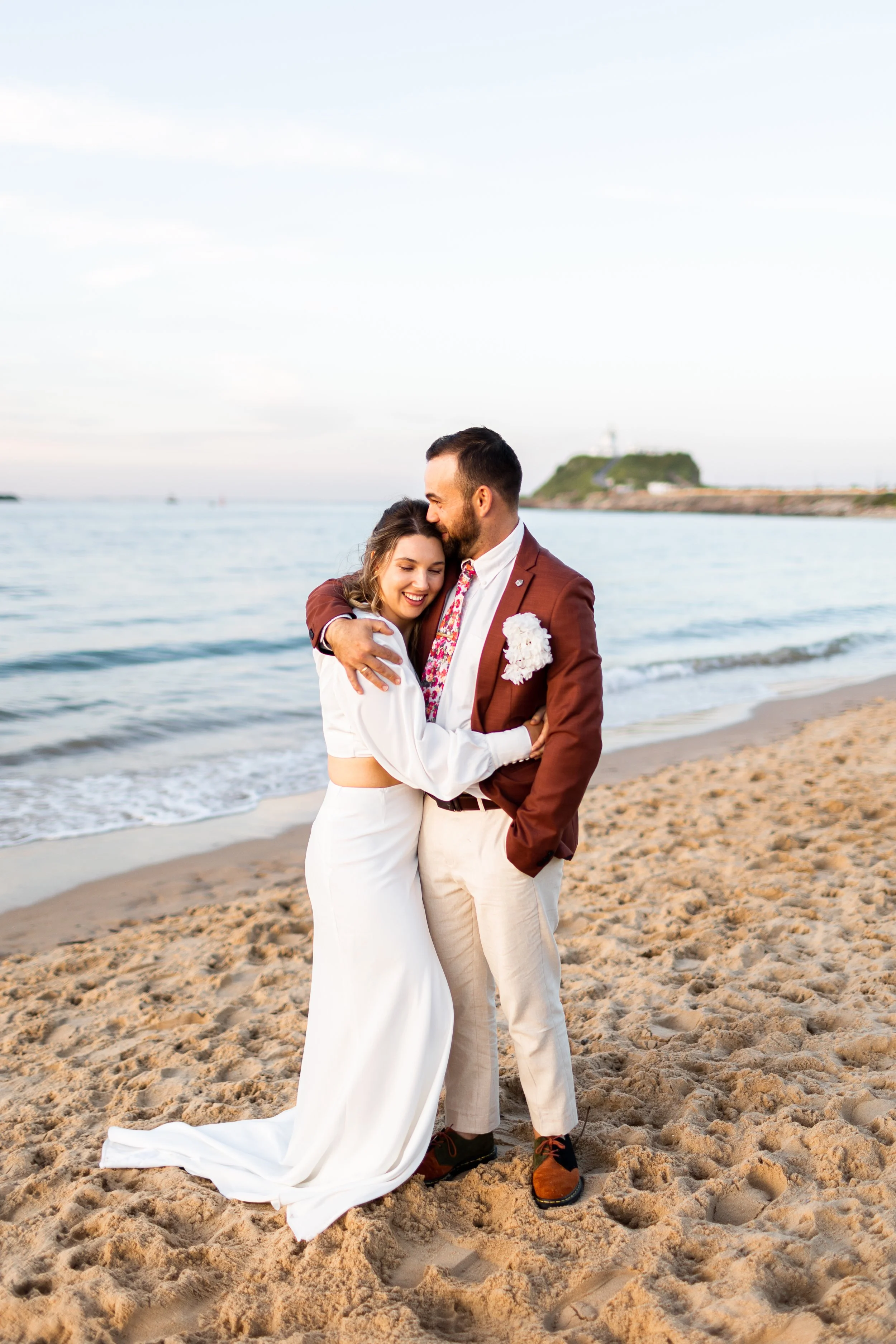A smiling couple hugging on the beach, dressed in wedding attire, with the ocean and a distant island in the background.