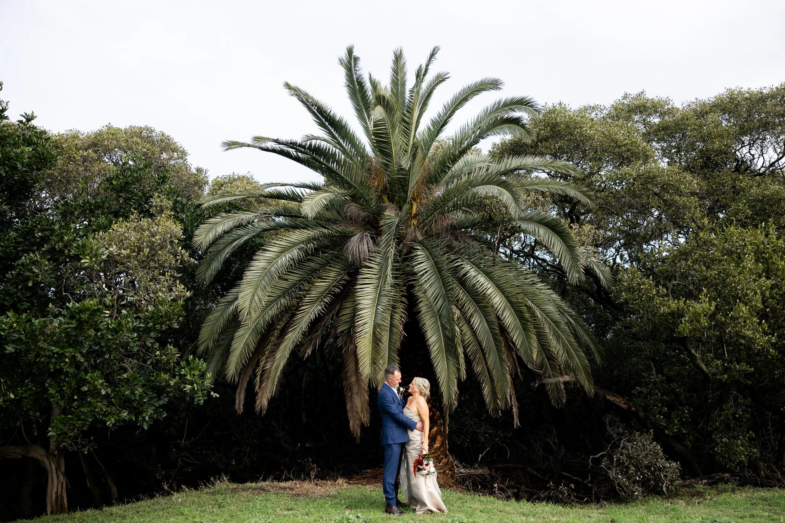 A newlywed couple standing in front of a large palm tree, holding hands and looking at each other, outdoors with greenery and cloudy sky in the background.