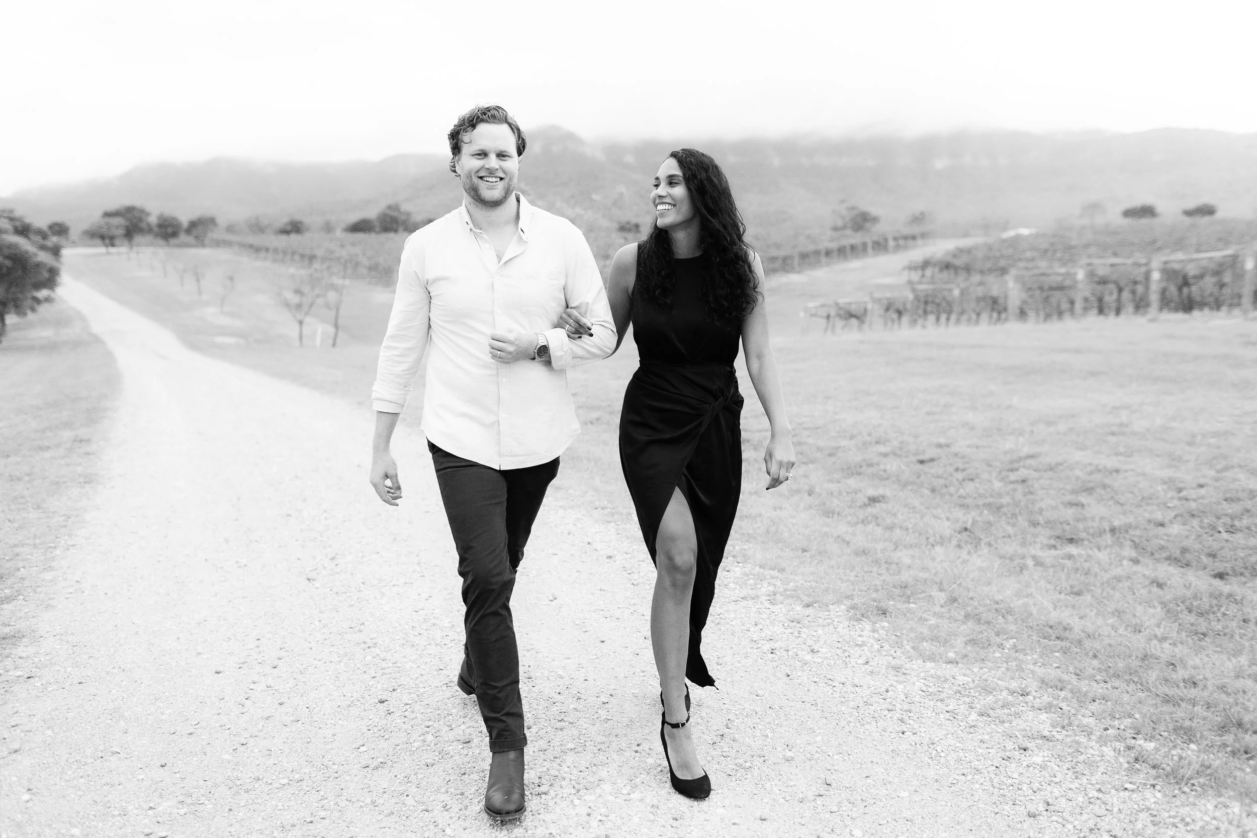 A man and woman walking arm in arm on a dirt road in a rural landscape, smiling and looking at each other.