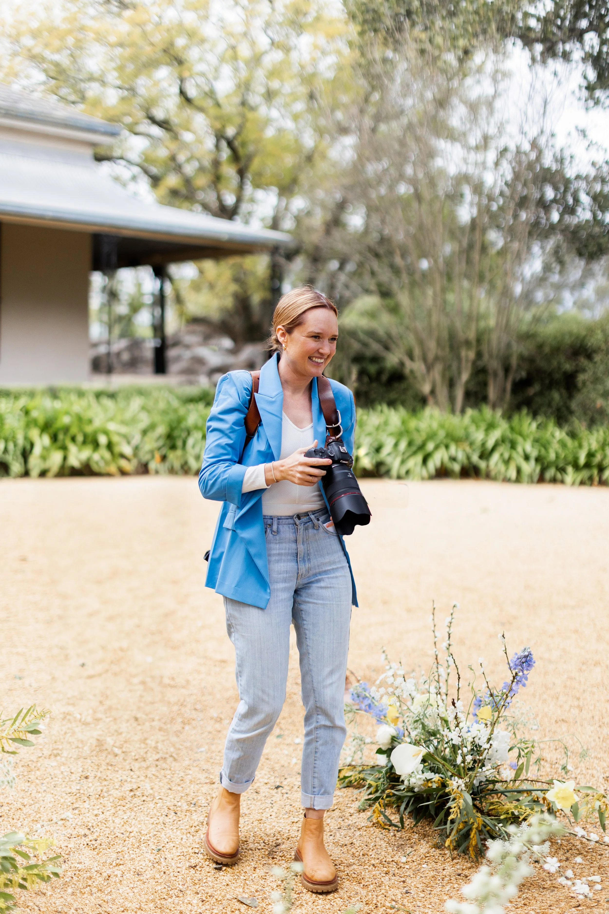 A woman holding a camera, smiling, standing outdoors near a floral arrangement
