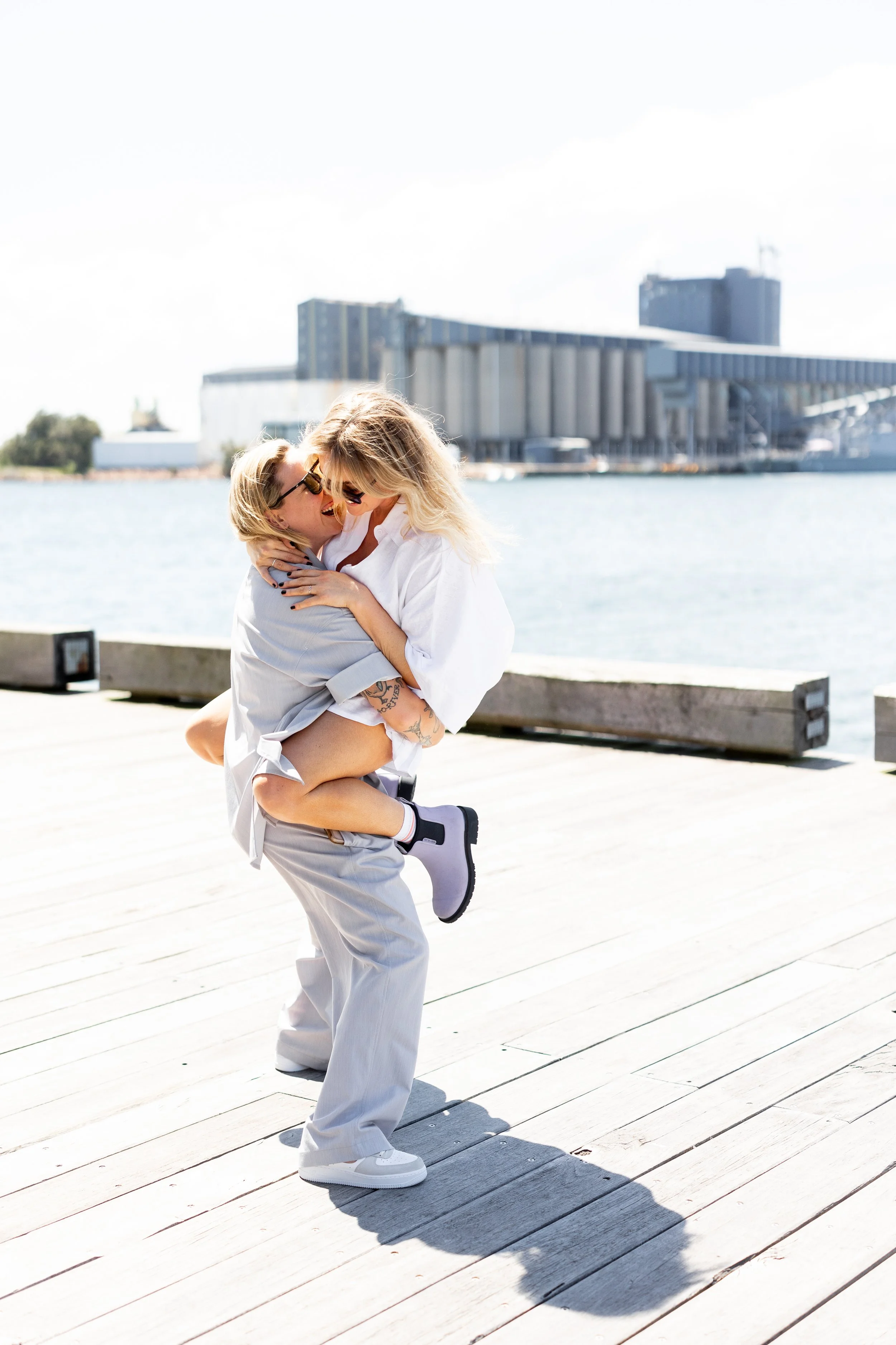 Two women are joyfully hugging and laughing on a wooden pier near the water with modern buildings in the background.