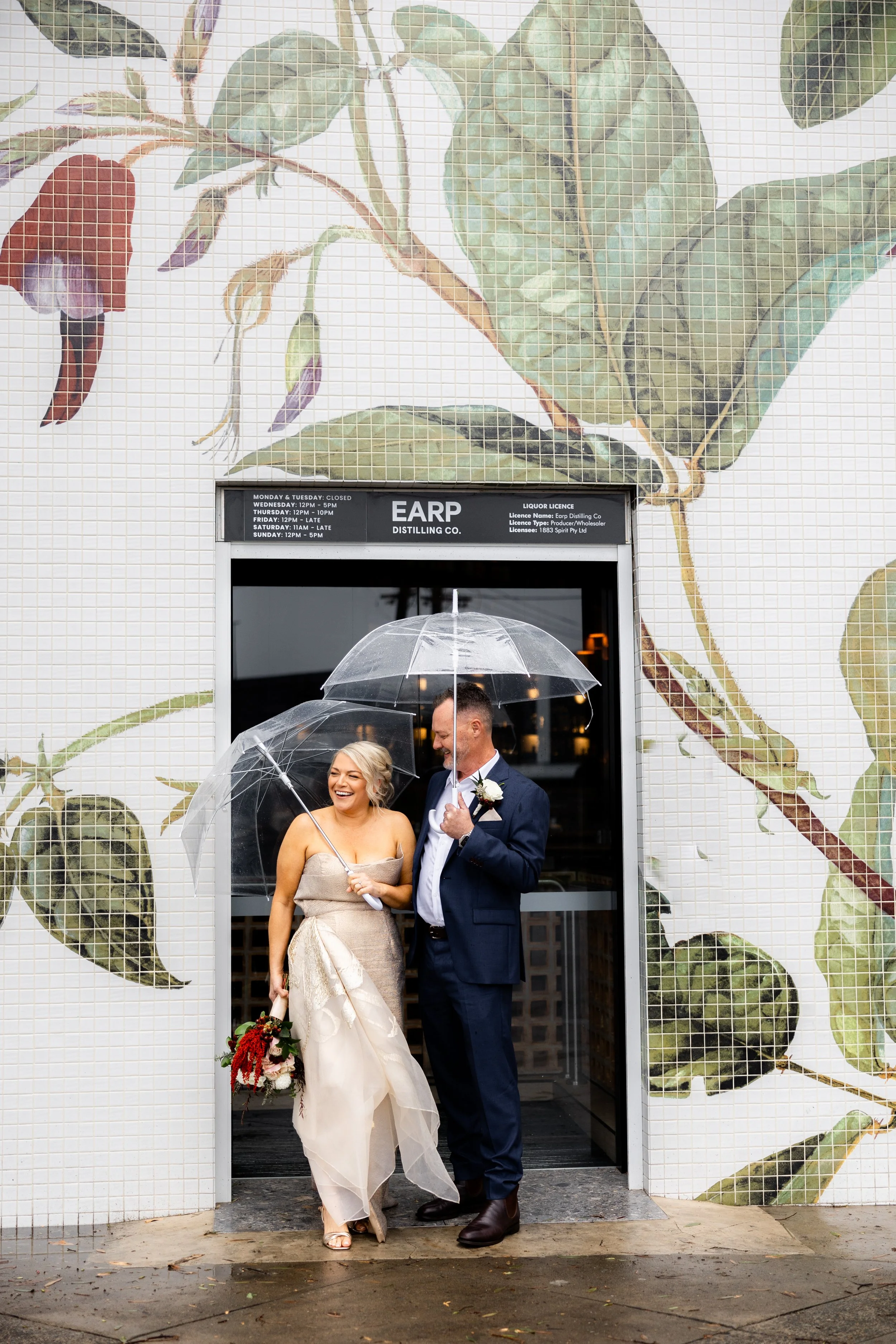 A smiling woman in a light-colored wedding dress holding a bouquet of flowers, and a man in a navy suit holding a clear umbrella, standing in front of a door with a large mural of green leaves on the wall behind them, holding a clear umbrella on a ra