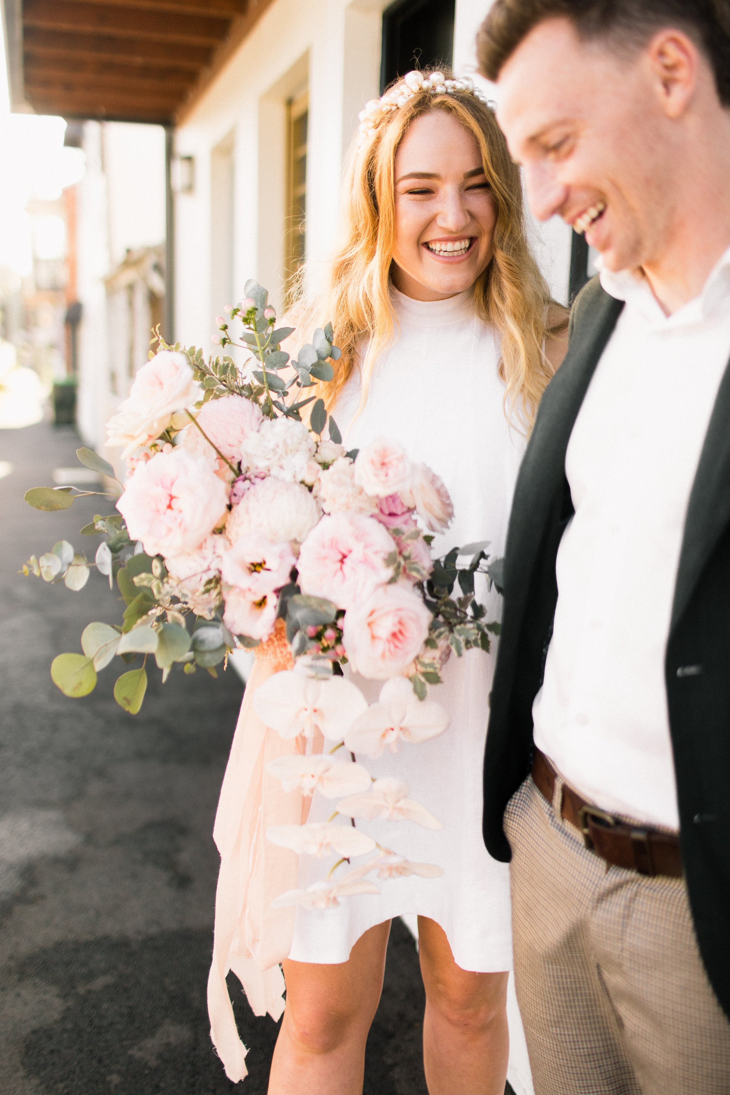 A woman smiling and wearing a flower crown holds a large bouquet of pink and white flowers, standing beside a man in a suit on a sunny street.