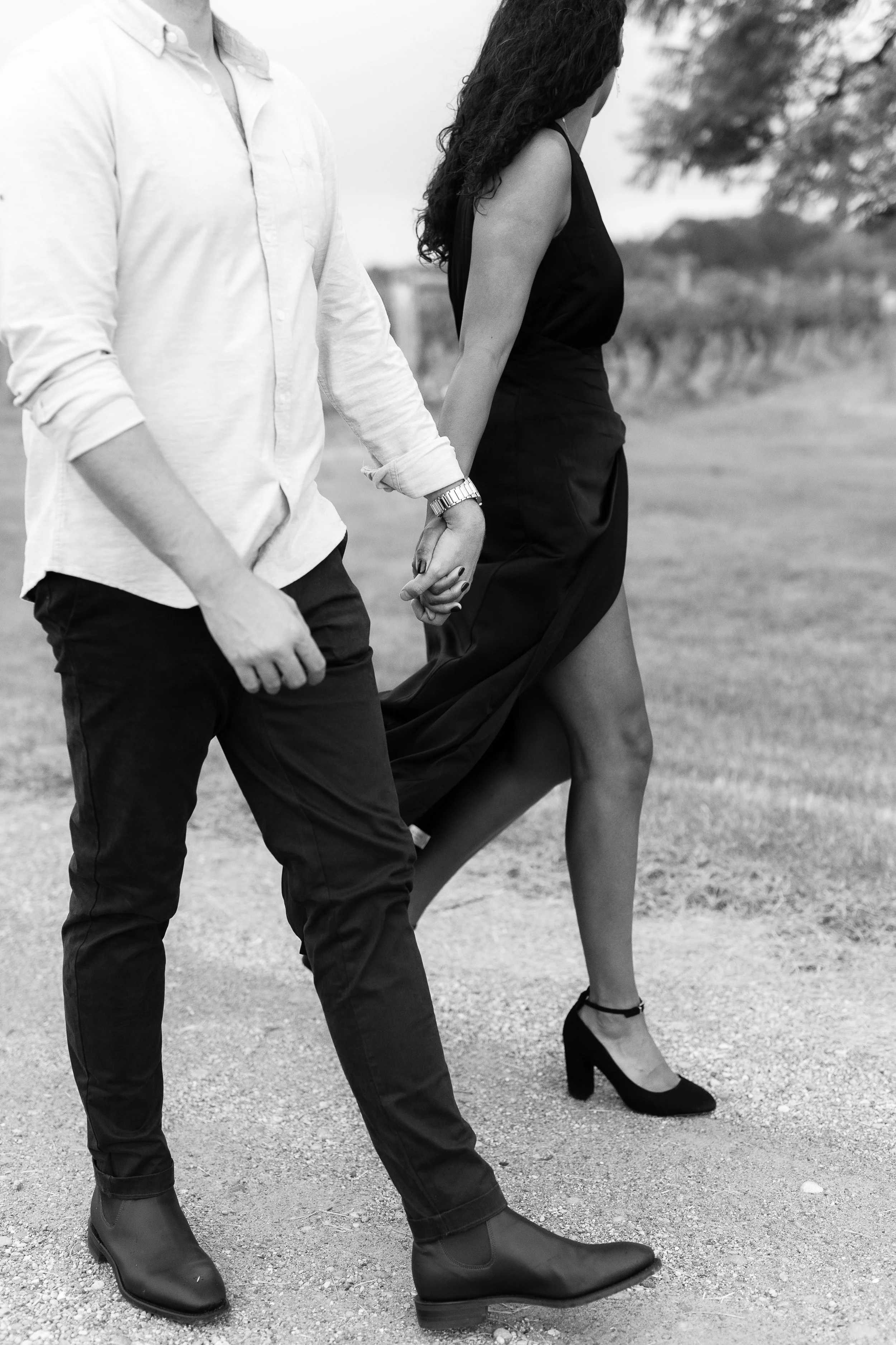 A black and white photograph of a man and woman holding hands outdoors. The woman is wearing a long black dress and high heels, while the man is wearing a white shirt, dark pants, and boots.