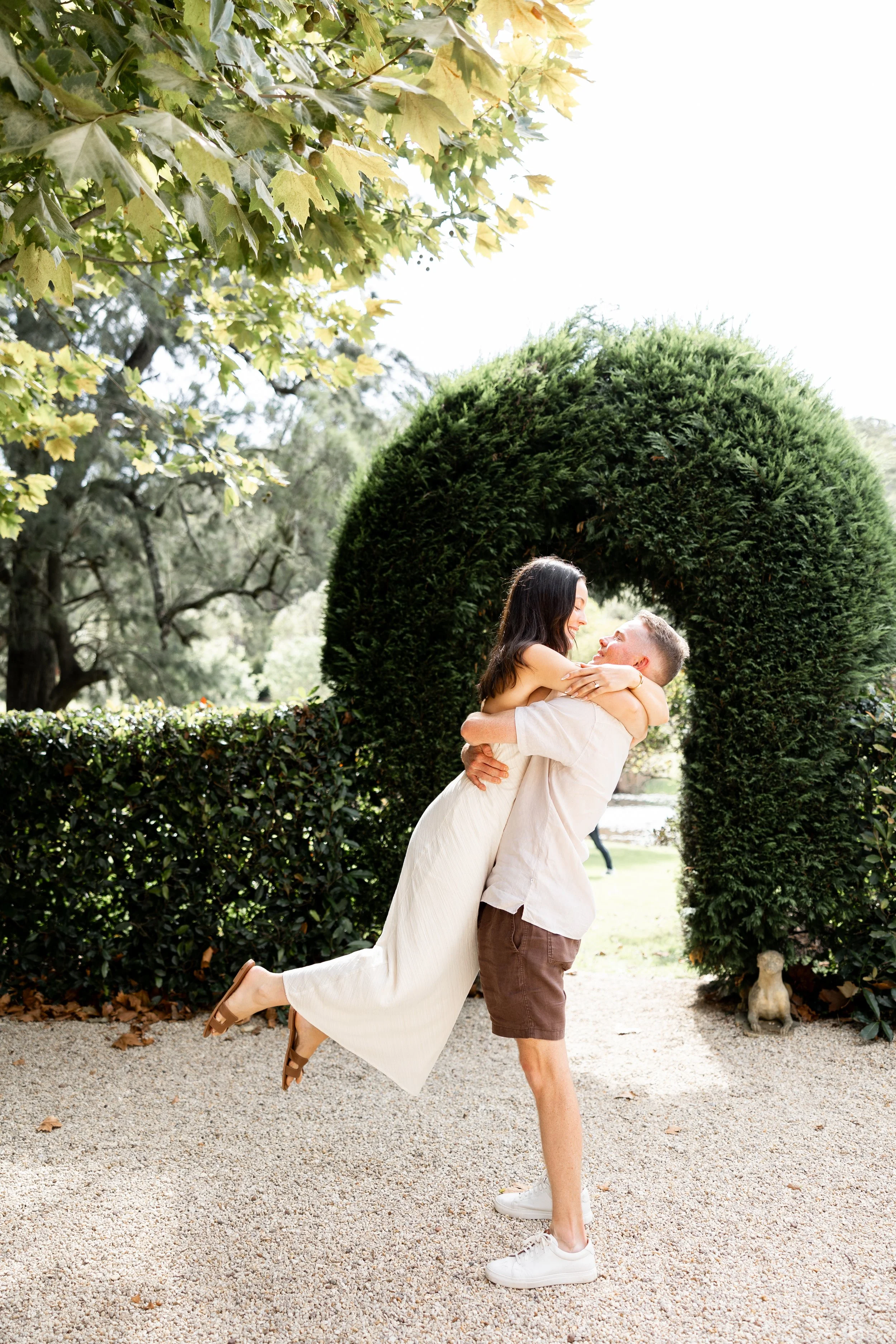 A man holding a woman who is smiling and looking at him, in an outdoor park setting with greenery and a large circular shrub in the background.