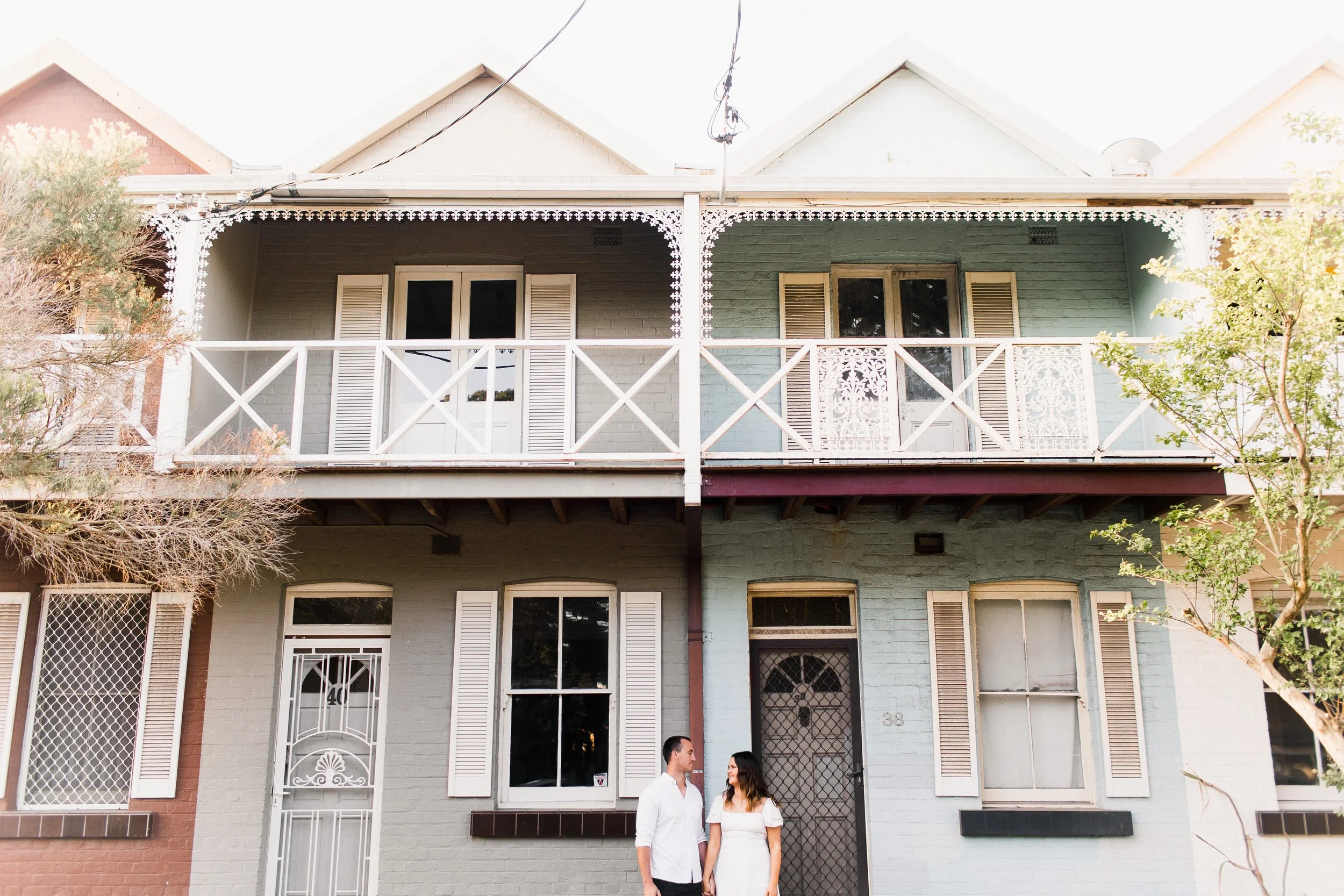 A man and woman standing in front of a two-story pastel-colored building with balcony and shuttered windows.