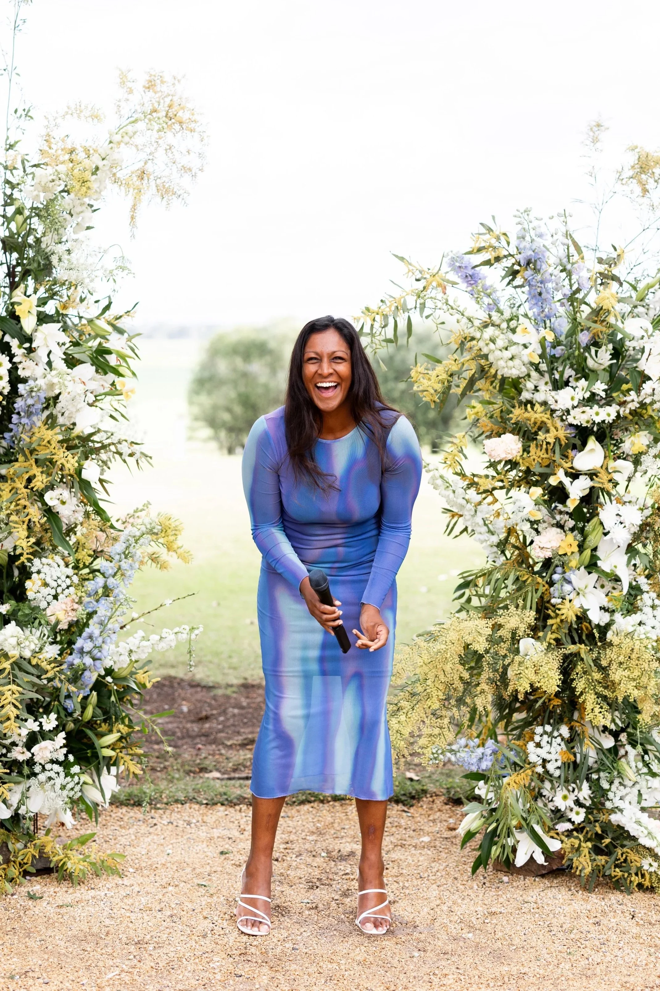 A woman in a blue dress holding a microphone, standing in front of floral arch at an outdoor event, smiling or laughing.