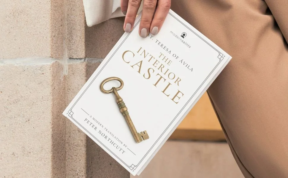 A person holds a book titled "The Interior Castle" by St. Teresa of Avila, with a golden key resting on the cover.