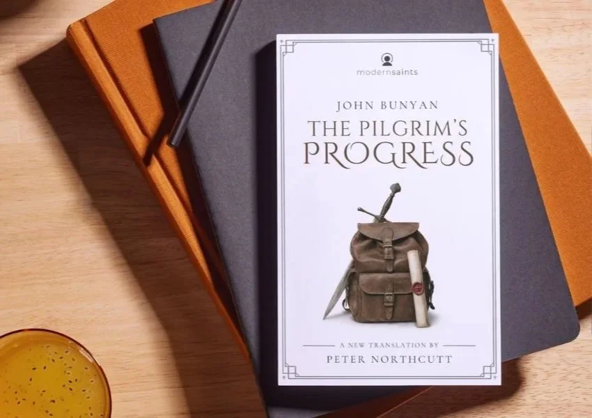 A book titled "The Pilgrim's Progress" by John Bunyan on top of a stack of books, with a pen resting on the stack, placed on a wooden surface near a glass of a light-colored beverage.