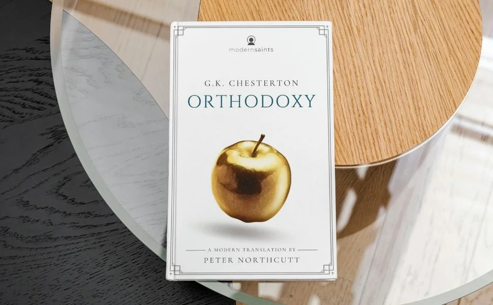 Book titled 'Orthodoxy' by G.K. Chesterton with a gold apple on the cover, placed on a glass table next to a wooden surface.