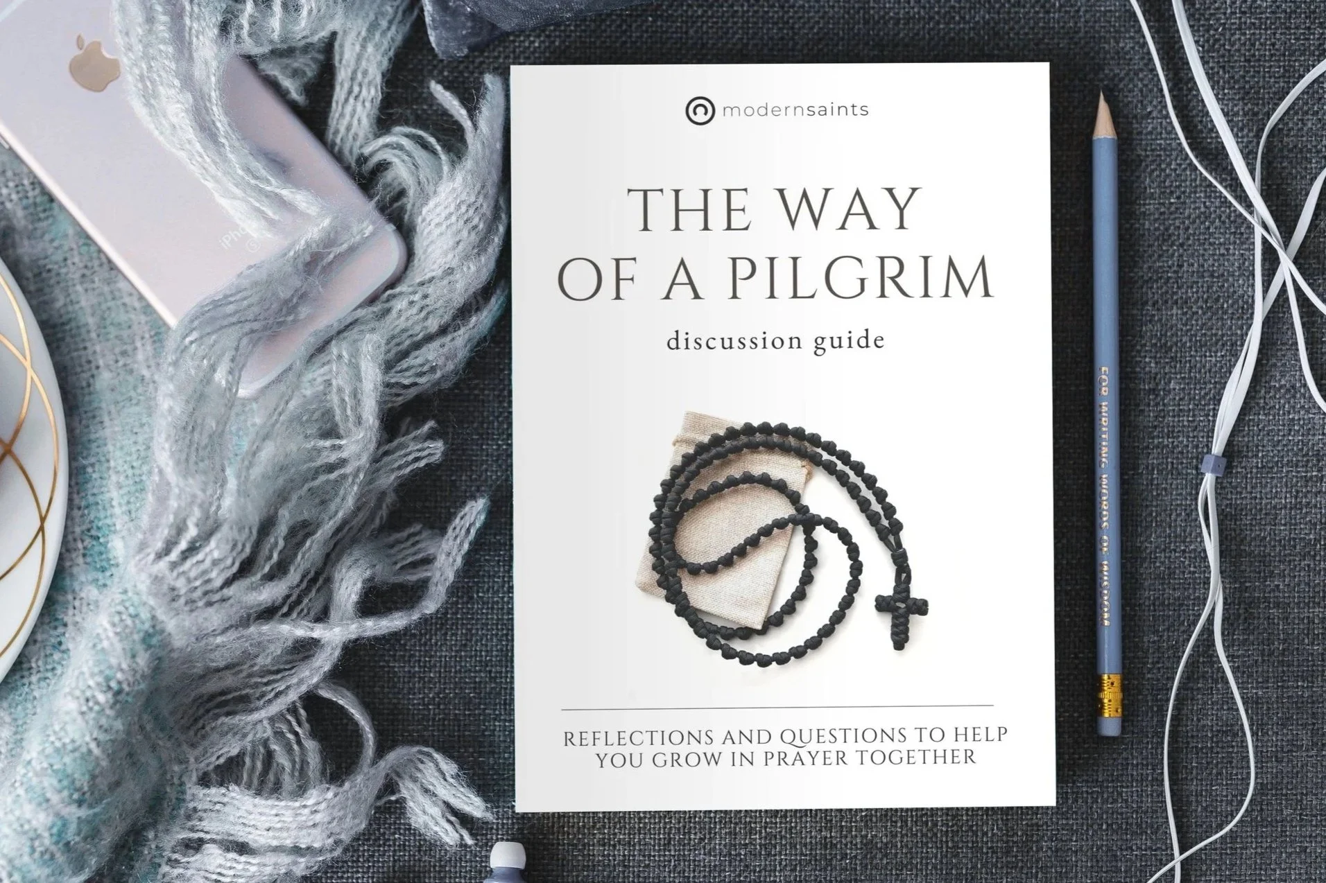 Discussion guide titled 'The Way of a Pilgrim' with prayer beads and a book on a gray cloth surface, accompanied by a gray scarf, a gold-rimmed plate, an Apple device, a blue pencil, and white earphones.