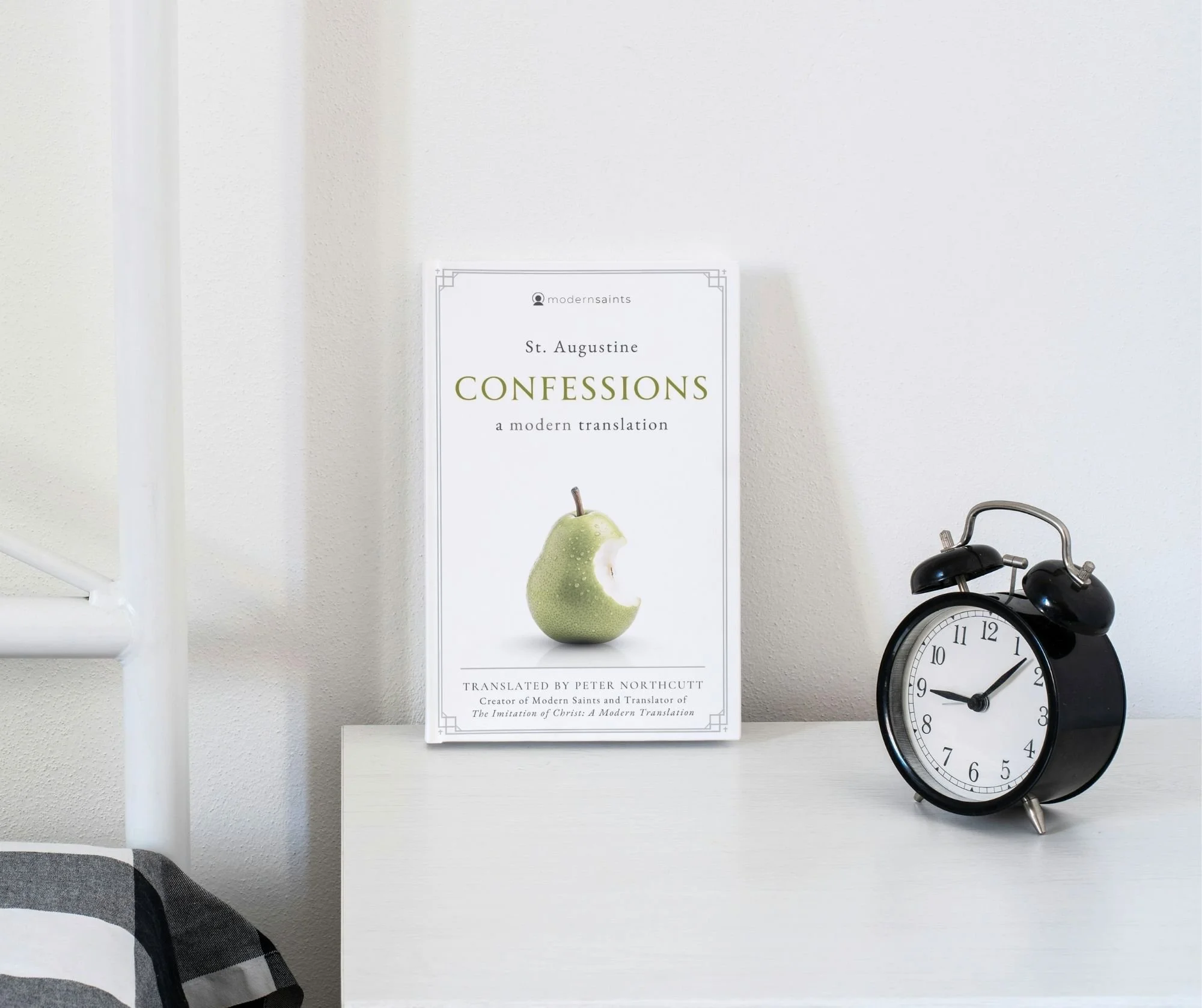 Book titled Confessions by St. Augustine with an image of a green apple with a bite taken out on the cover, placed on a white surface next to a black alarm clock showing 9:10, against a light-colored wall.