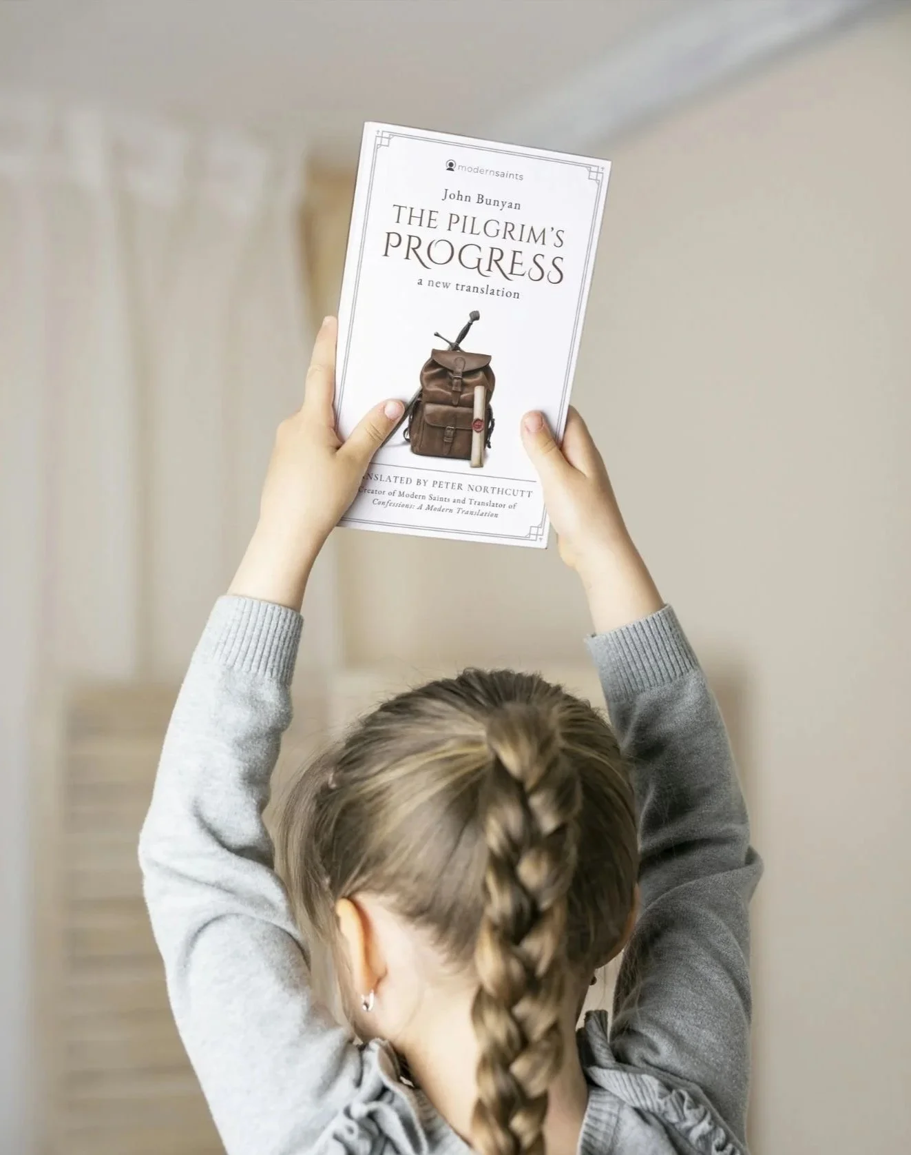 A young girl holds a copy of the book 'The Pilgrim's Progress' by John Bunyan above her head with both hands, showing the front cover.