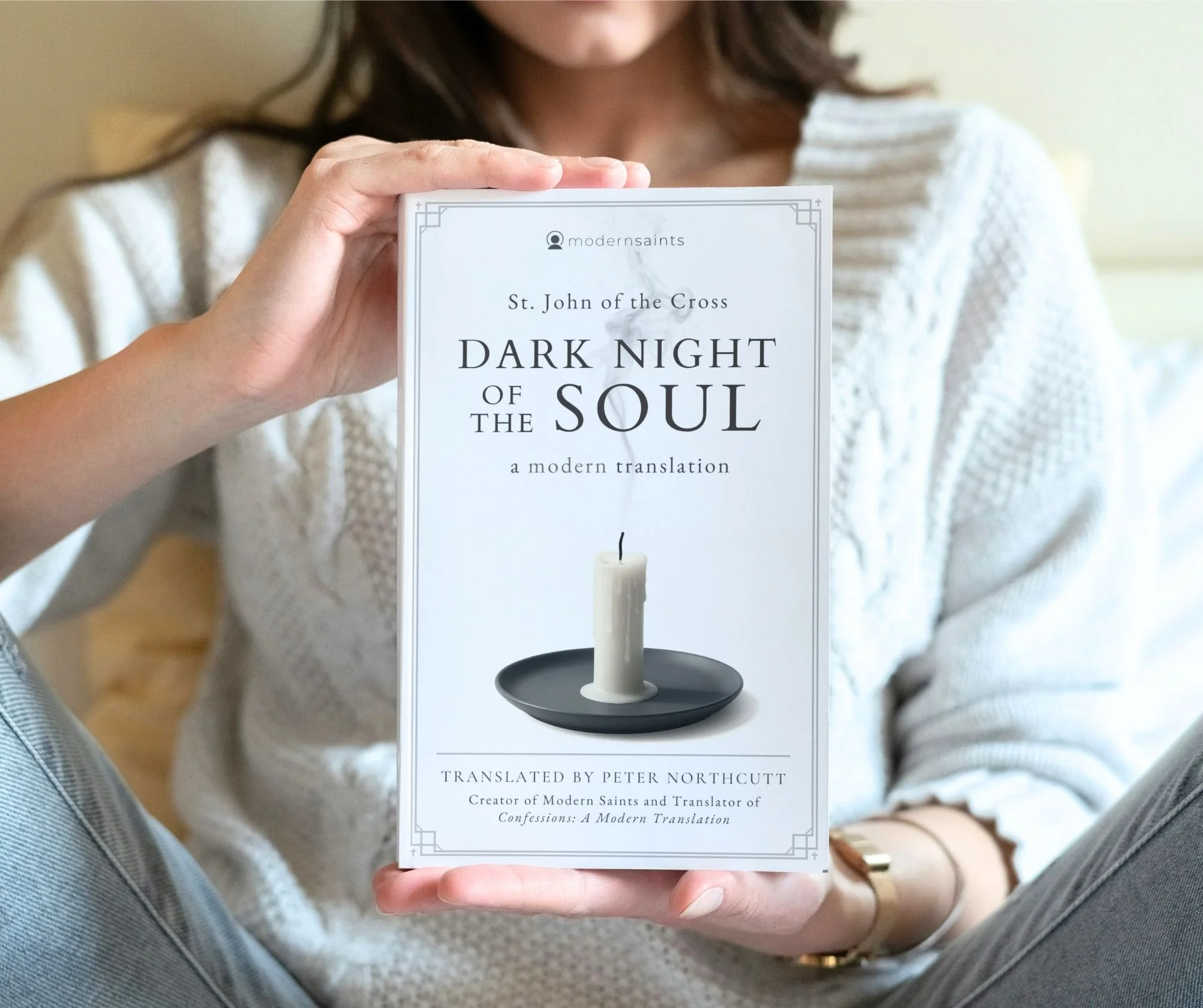 A person in a white sweater holding a book titled 'Dark Night of the Soul' by St. John of the Cross, translated by Peter Northcutt, with a white candle on a black dish on the cover.