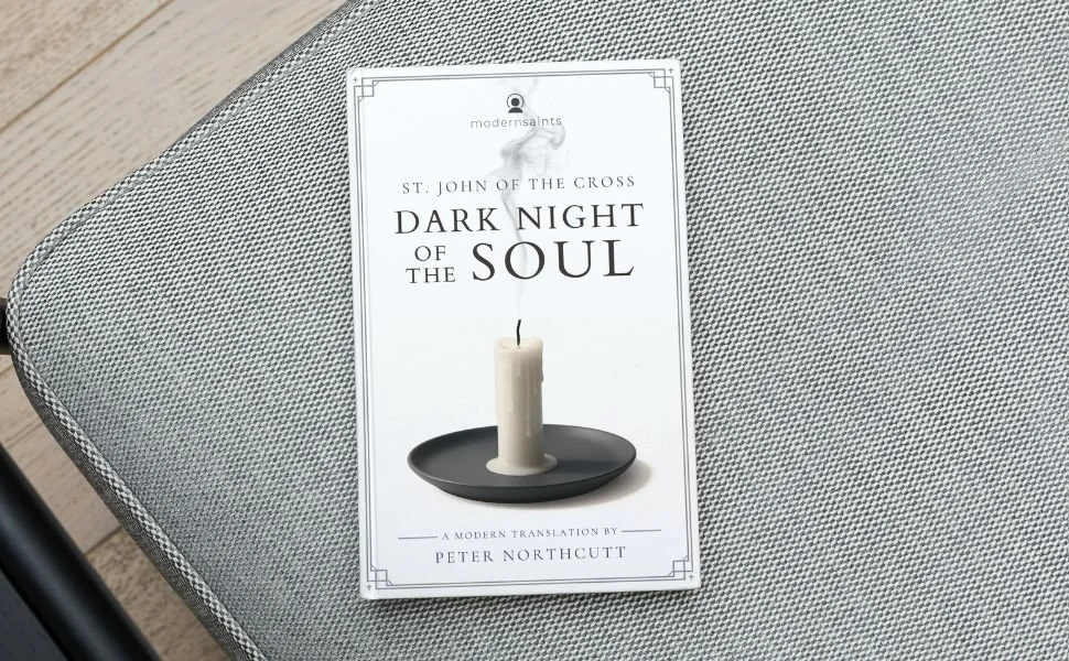 A book titled "Dark Night of the Soul" by St. John of the Cross, translated by Peter Northcutt, is placed on a gray textured fabric surface.
