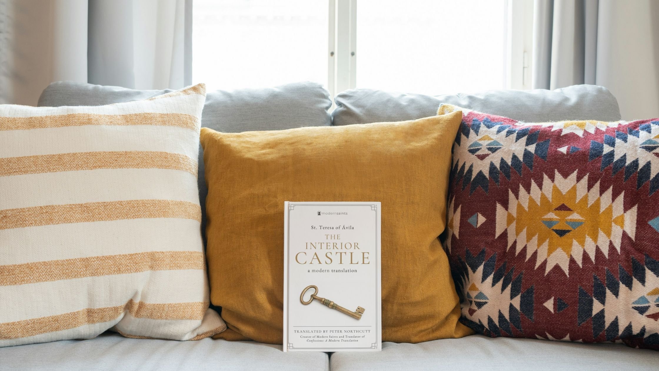 Three decorative pillows on a couch with a book titled 'The Interior Castle' by St. Teresa of Ávila resting against the pillows.