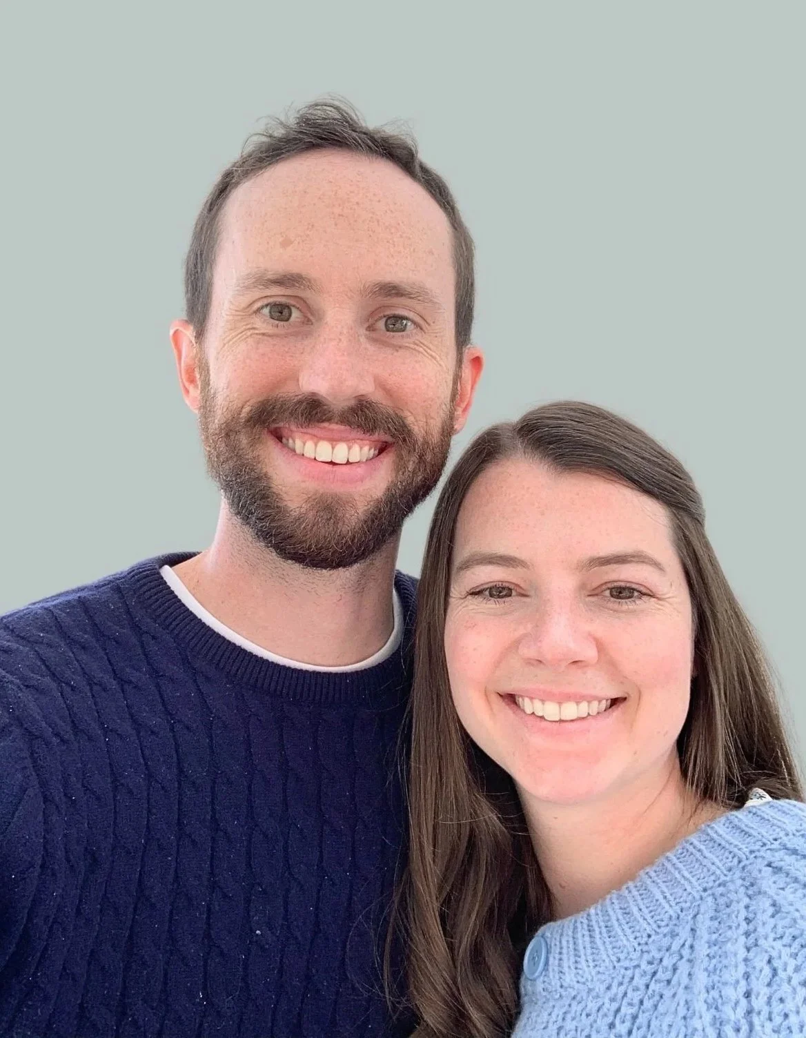 A smiling man with a beard and a woman with long brown hair, both wearing sweaters, standing close together against a plain background.