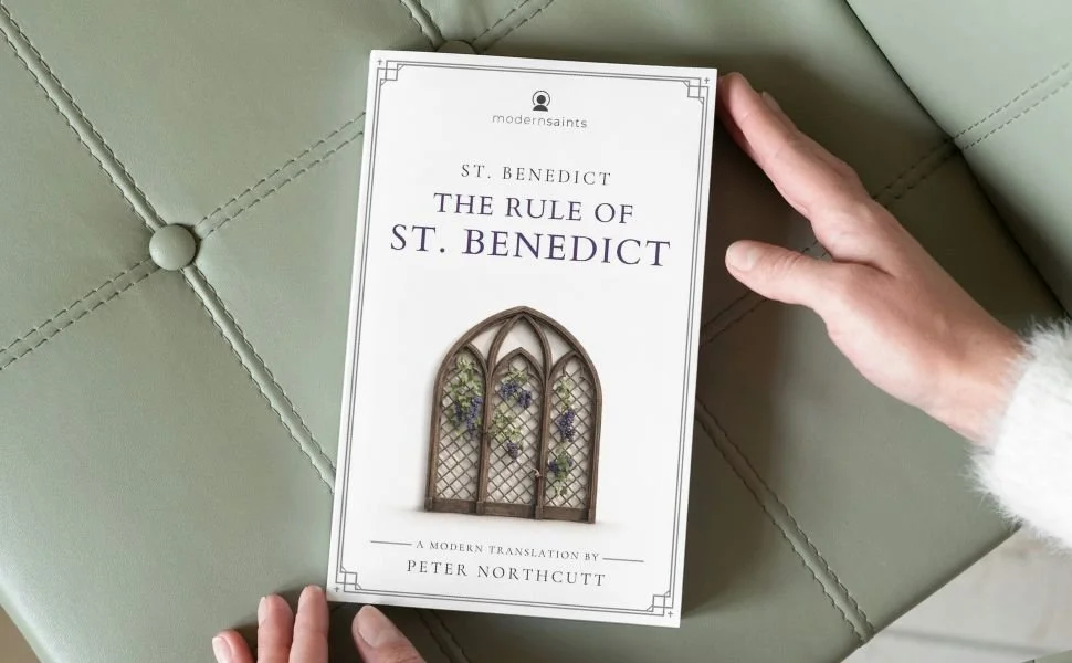 Person holding a book titled 'The Rule of St. Benedict a modern translation' by St. Benedict, with an illustration of a stained-glass window on the cover, on a green upholstered surface.
