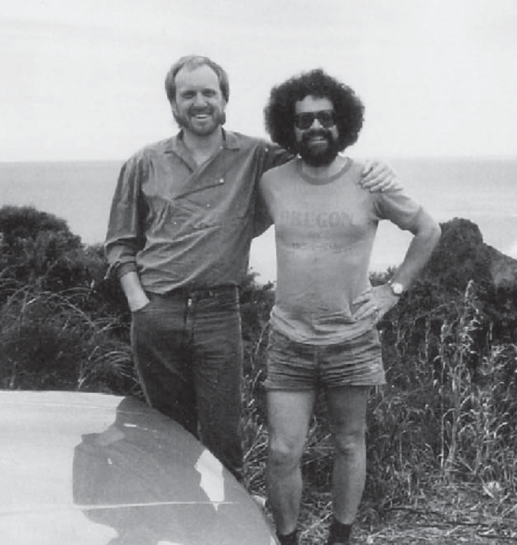 Deux hommes souriants debout sur une plage ou un rivage, avec des buissons derrière eux, l'un portant une chemise à manches longues et l'autre un t-shirt avec des rayures et des shorts. L'homme à droite porte des lunettes de soleil et une montre.