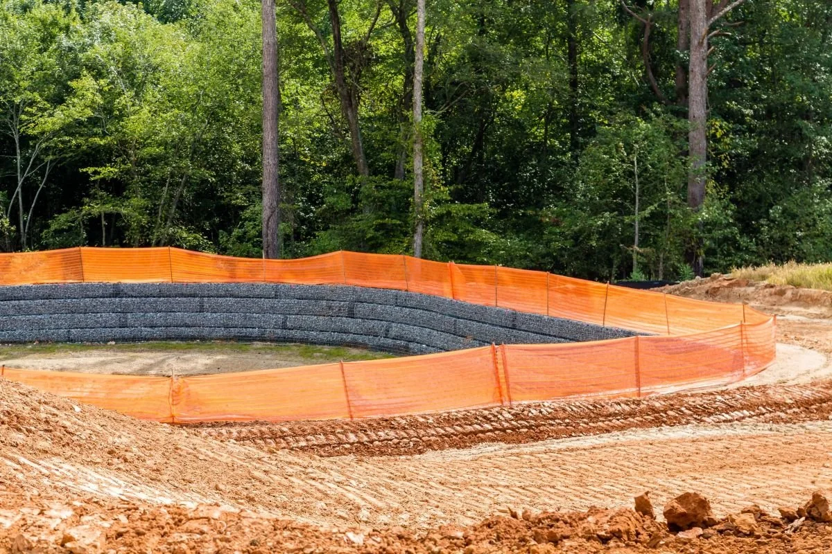 Construction site with orange safety fencing surrounding a curved area, black gravel wall, and dirt ground, with a background of green trees and foliage.