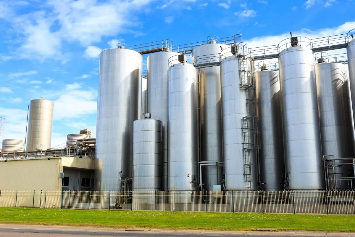 Tall silver industrial storage tanks in an open facility with blue sky and some clouds overhead.