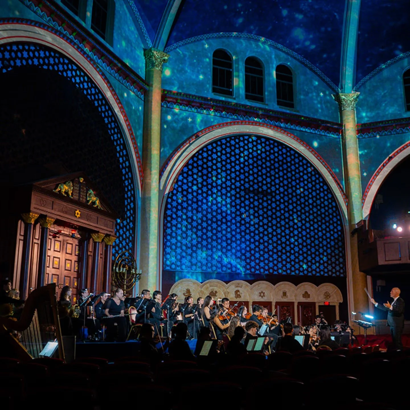 Orchestra and conductor under blue star-like projections at Temple Emanu-El