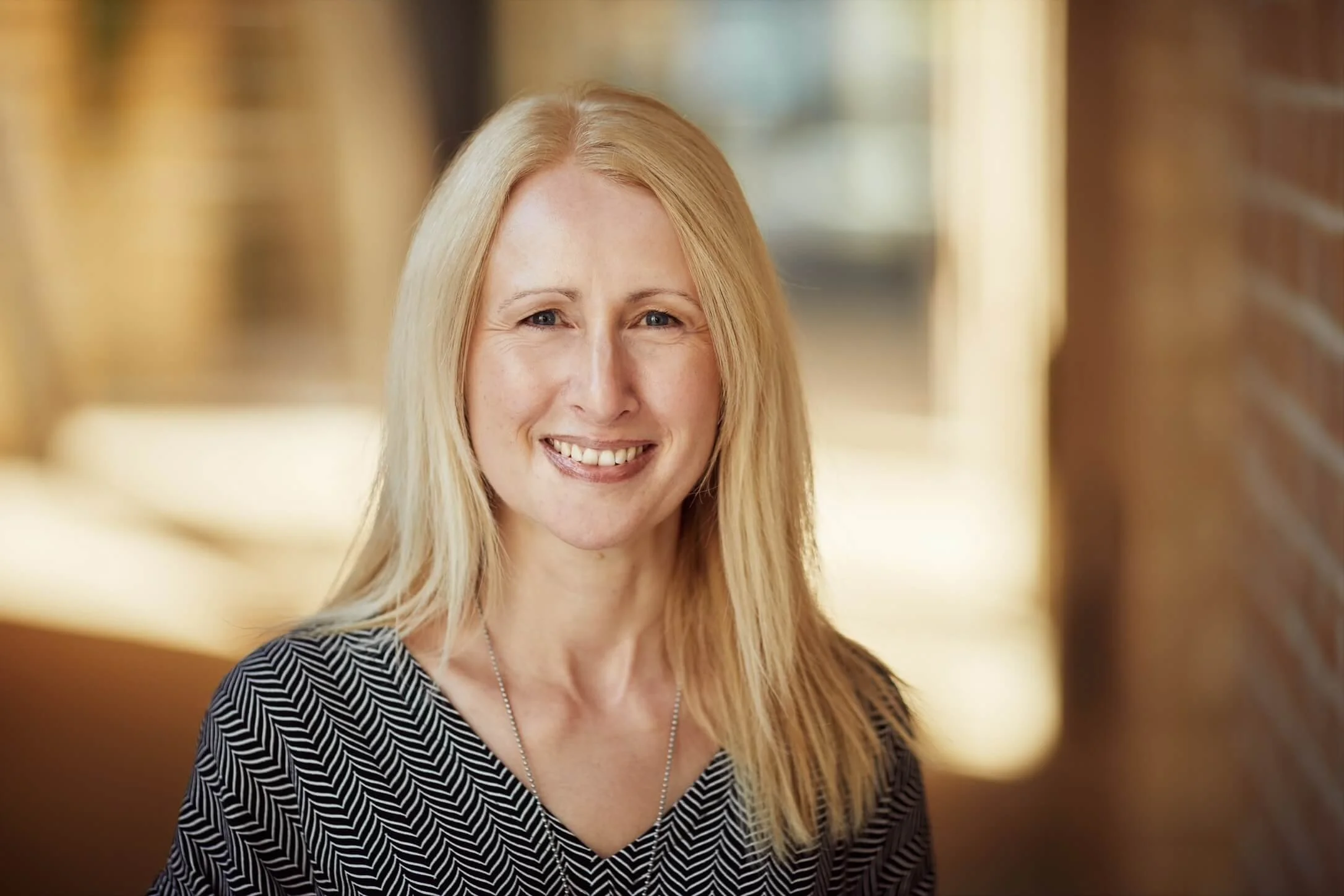 Smiling person with long blonde hair in a zigzag-patterned top, blurred outdoor background.