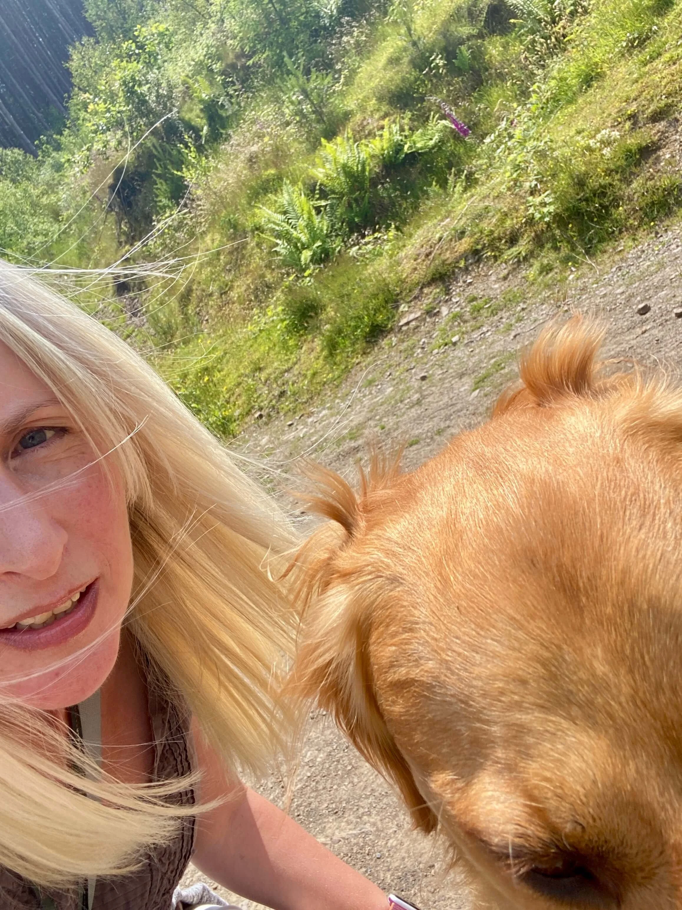Woman with blonde hair and a golden retriever outdoors on a trail with greenery in the background.
