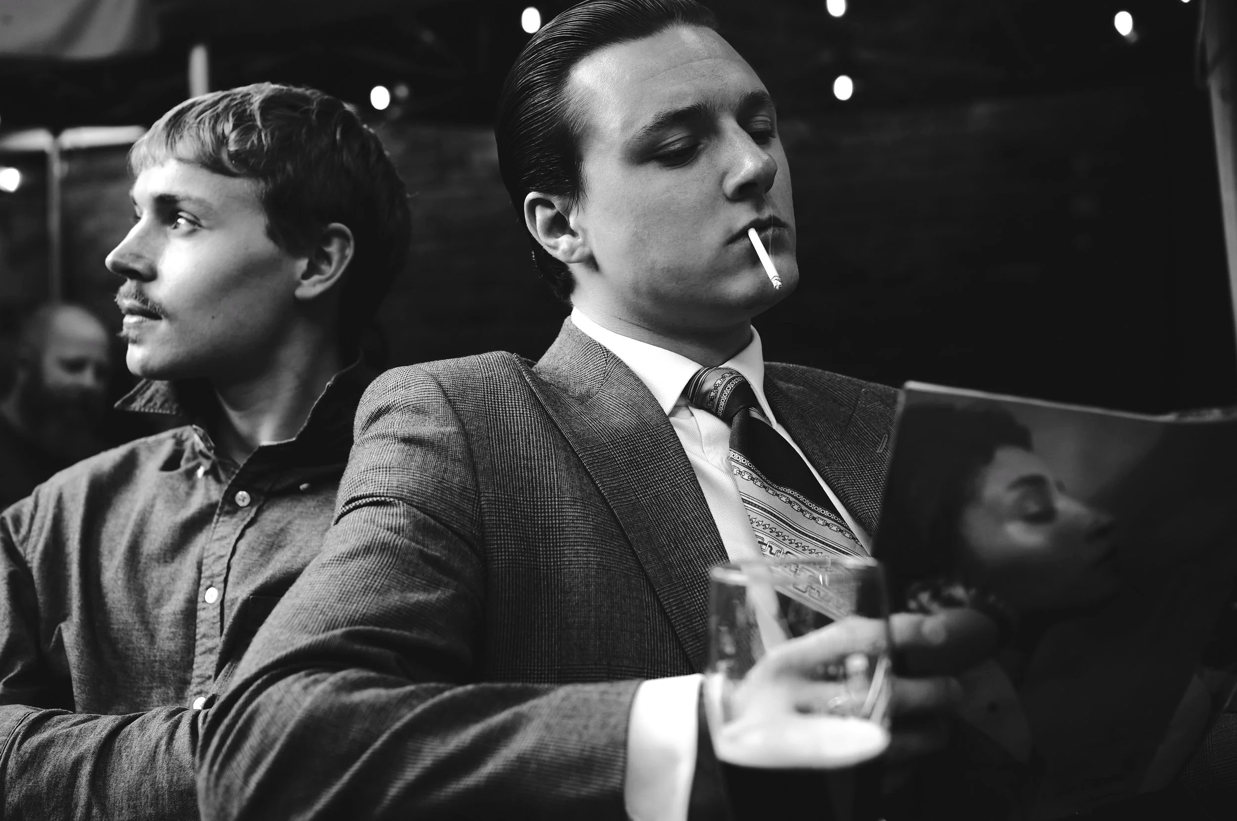 Two men sitting closely; one with a mustache facing left, and another in a suit smoking a cigarette and holding a drink. Black and white image.