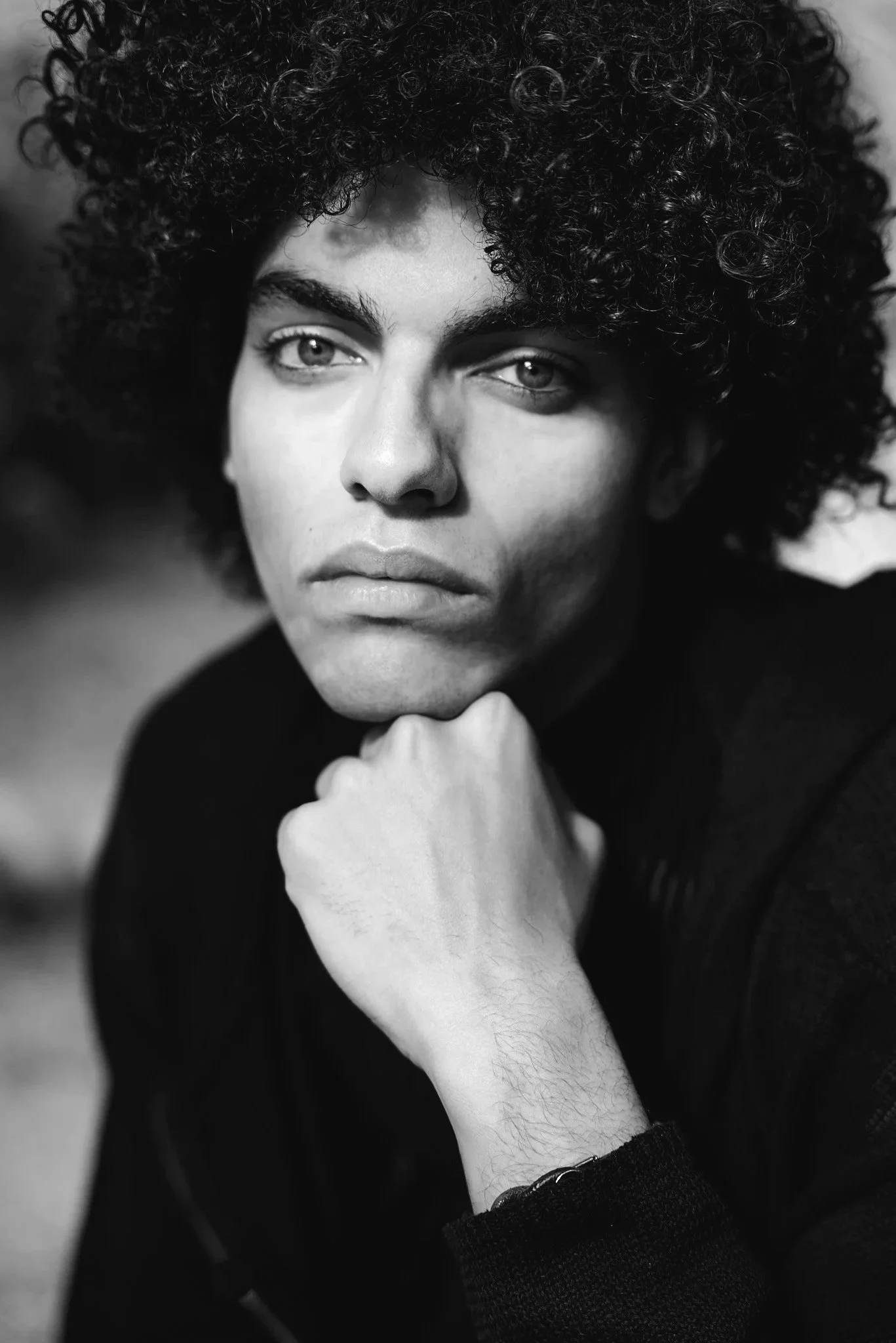 Black and white portrait of a person with curly hair looking directly at the camera, resting their chin on their fist.