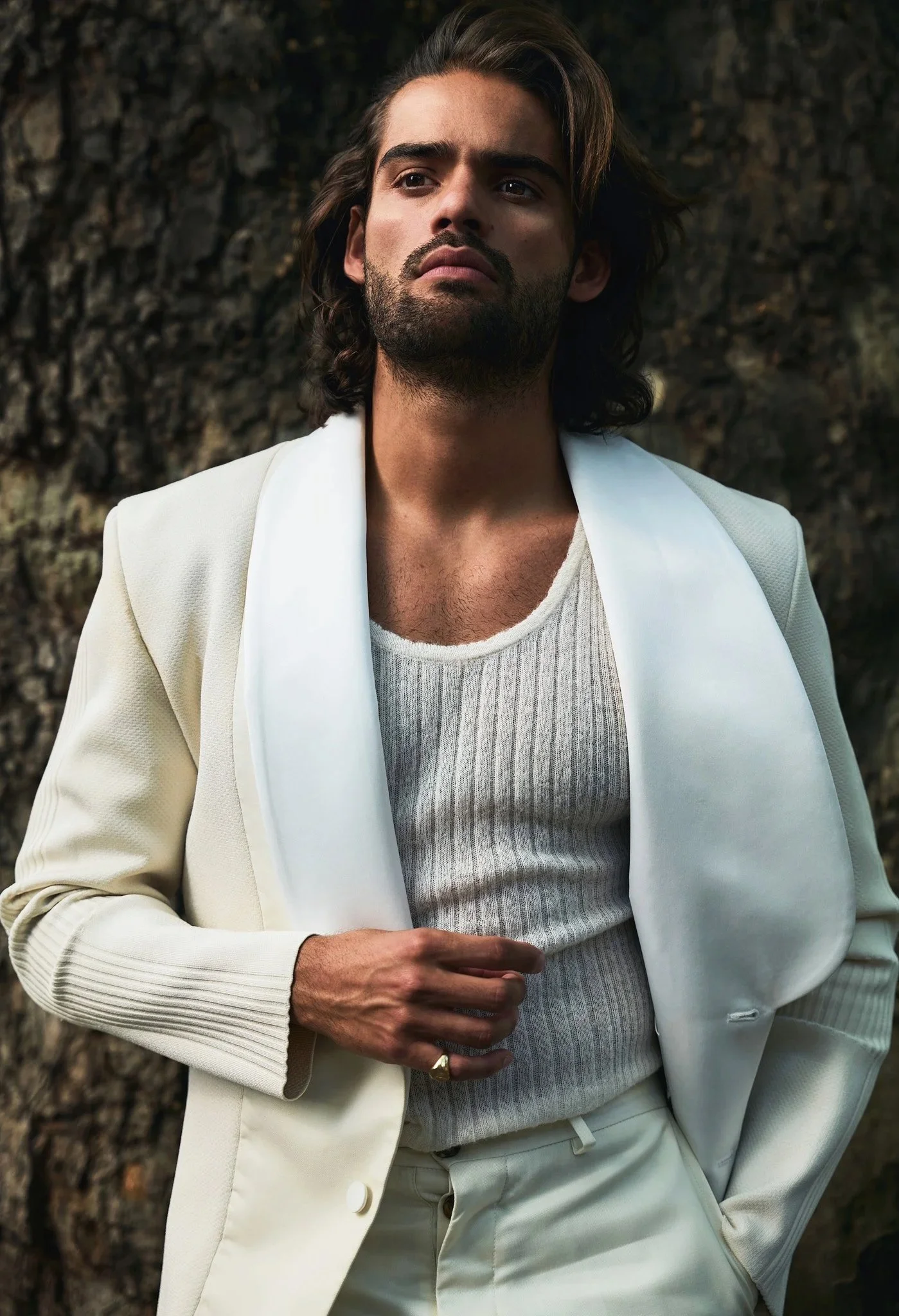 Man in cream suit and knit tank top standing outdoors with a textured tree bark background.