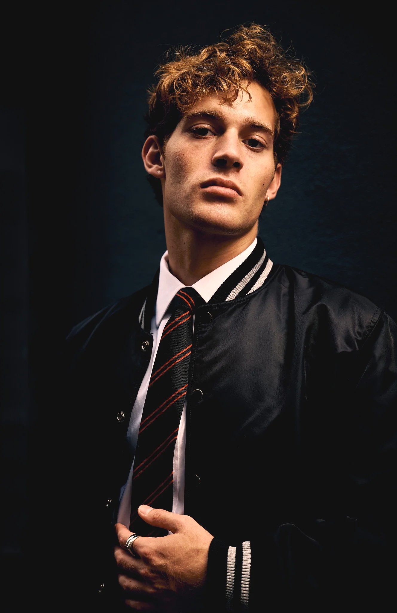 Portrait of a man with curly hair wearing a black jacket, white shirt, and striped tie, looking serious.