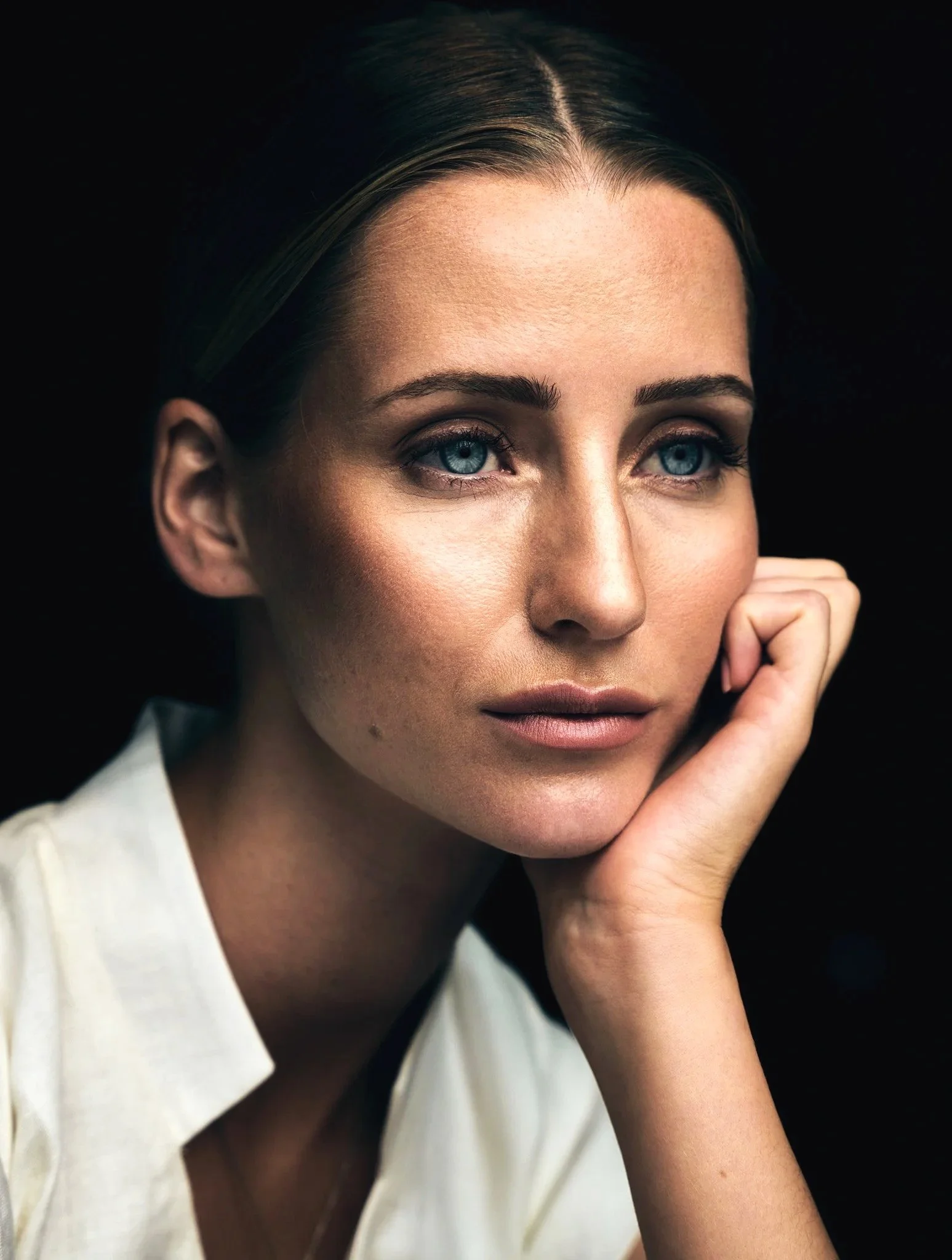 Close-up of a woman with blue eyes resting her chin on her hand, wearing a white shirt against a dark background.
