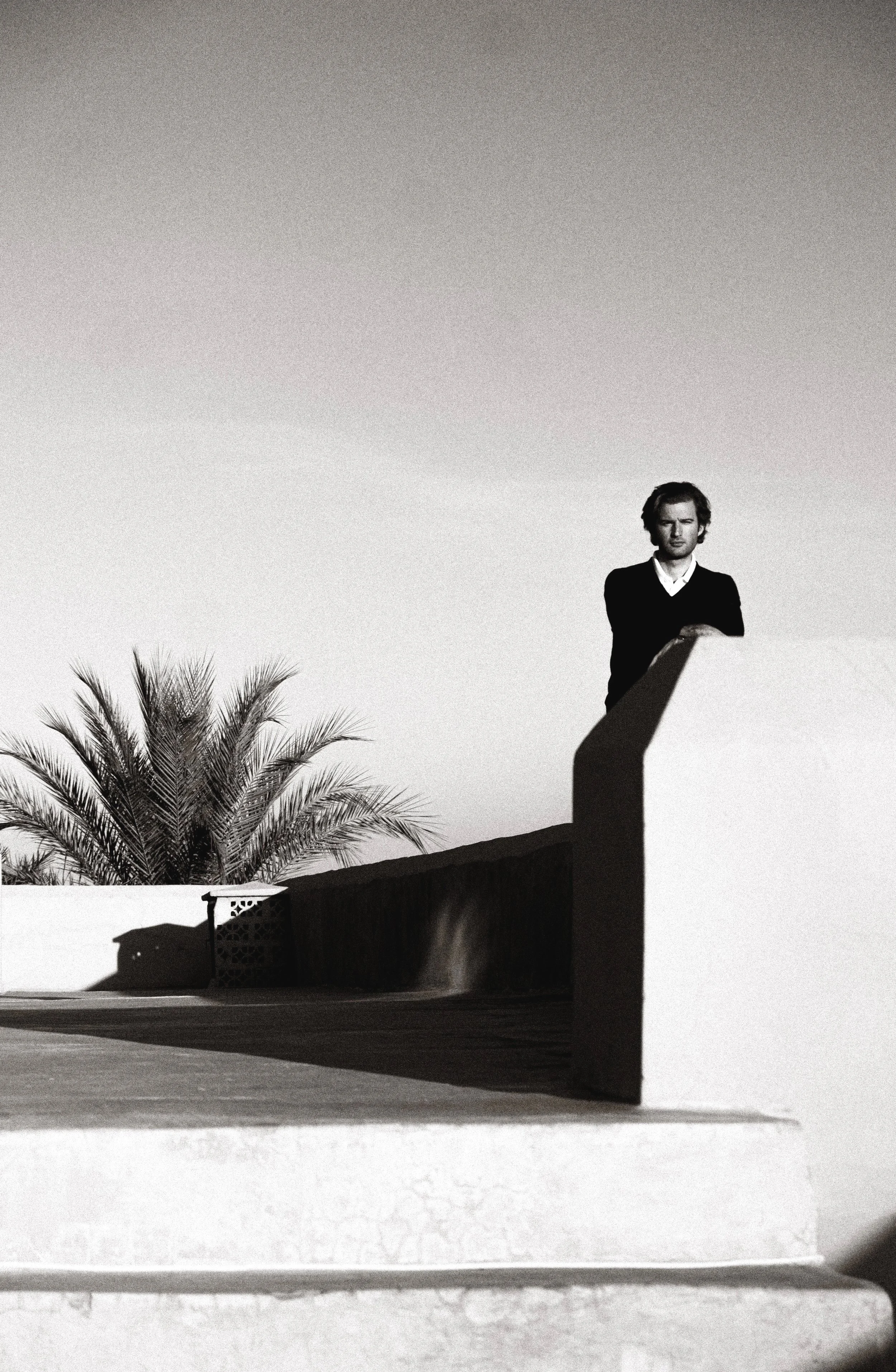 A black and white photograph of a person standing on a rooftop next to a palm tree, with a clear sky in the background. They appear contemplative, wearing a dark sweater over a light shirt.