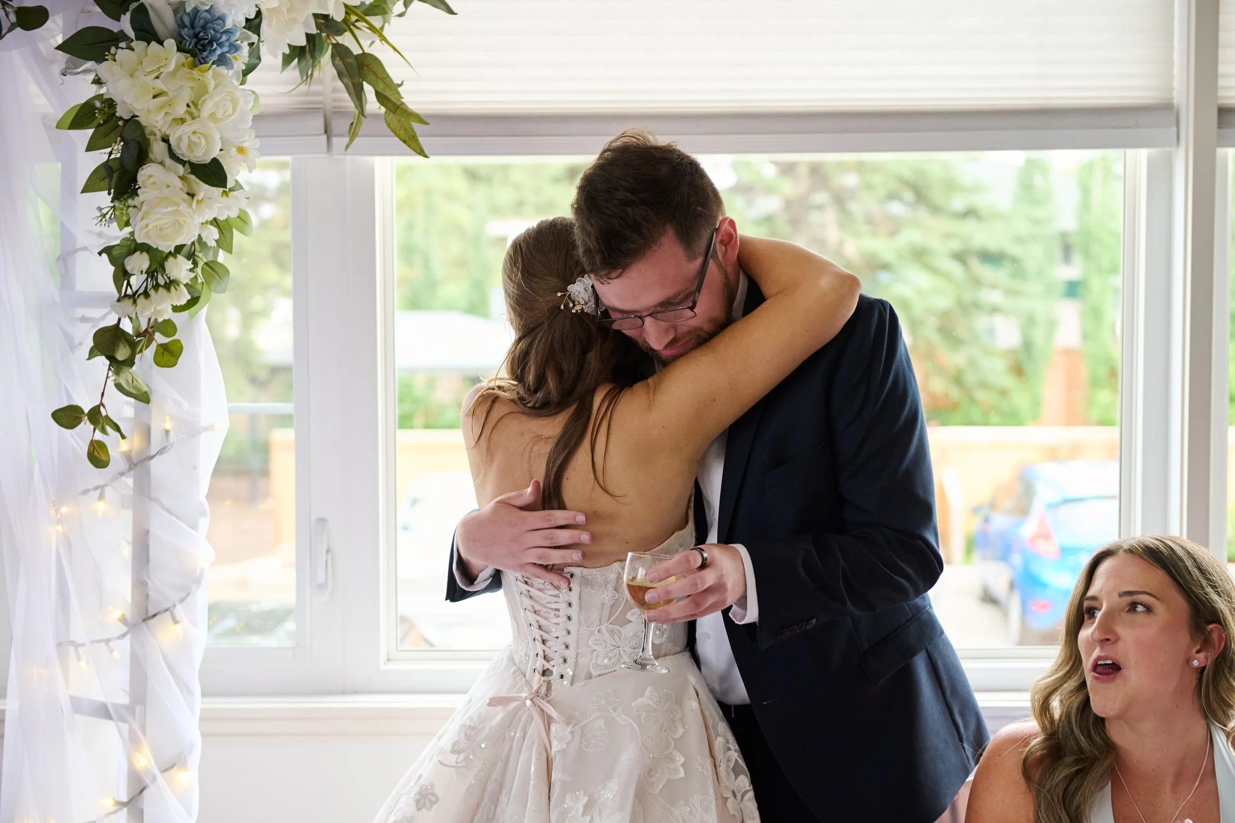 A bride and groom hug during their wedding celebration, with a bridesmaid sitting nearby, inside a decorated venue with flower arrangements and large windows.
