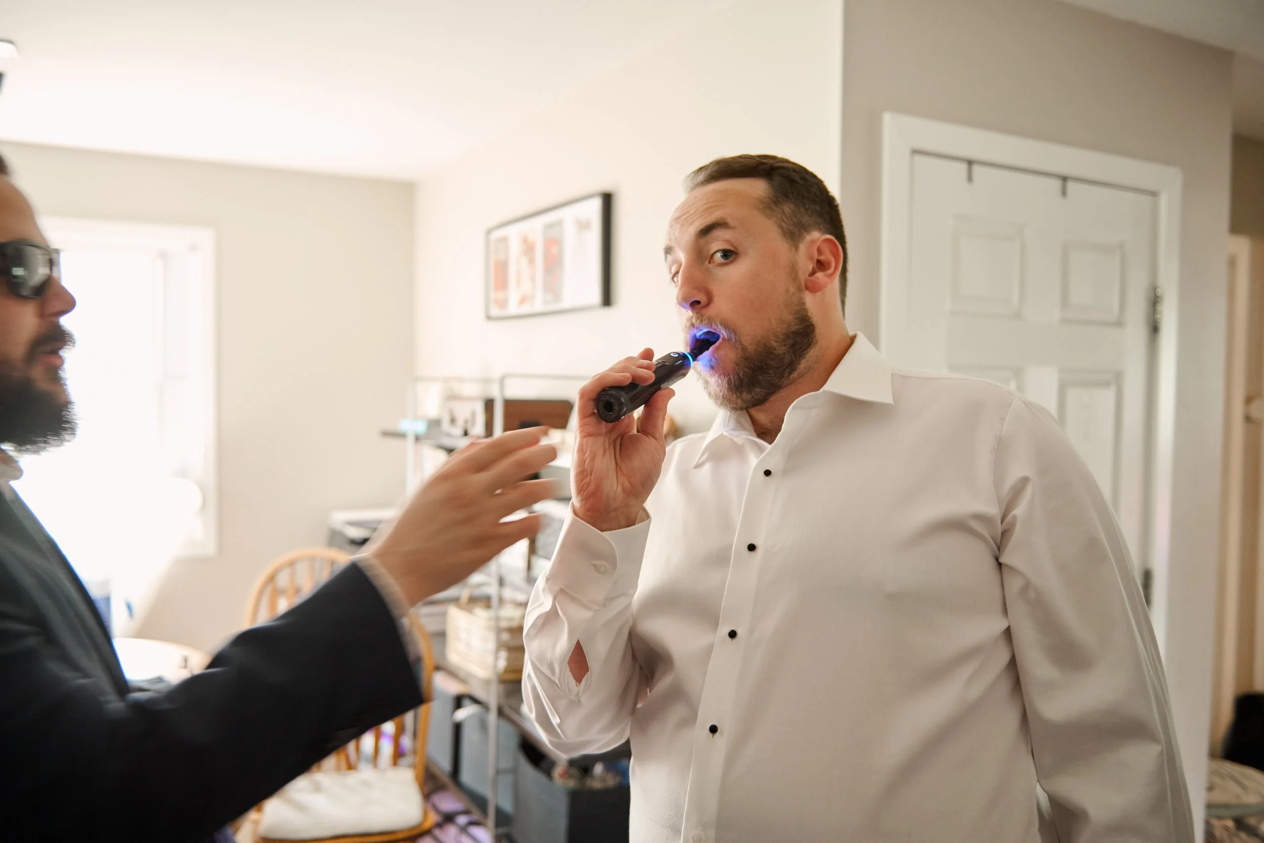 Man in white dress shirt using a black electric toothbrush to his mouth, with another person in a dark suit holding his hand, inside a home.