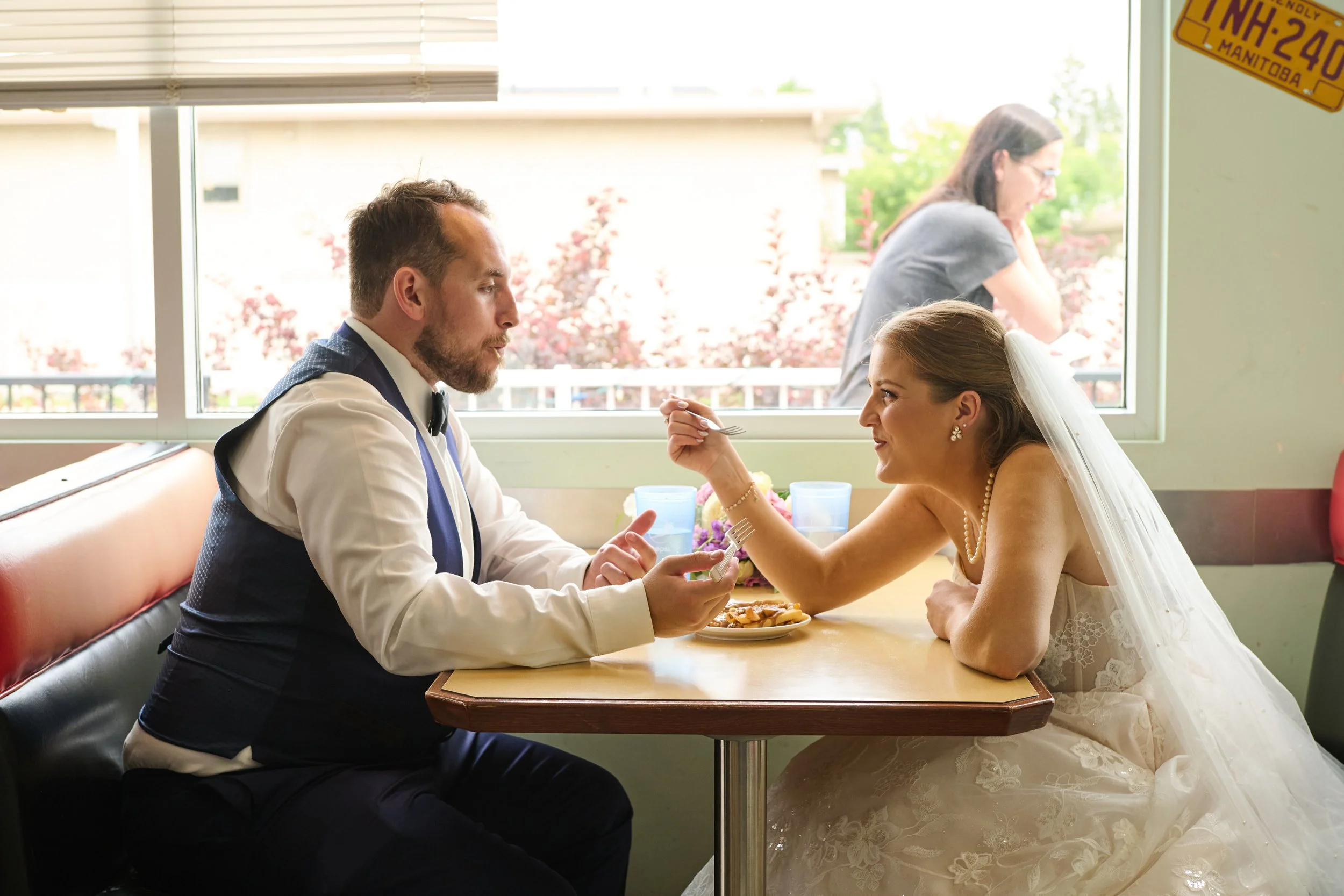 A bride and groom sit at a table inside a restaurant, the bride playfully feeding the groom with a fork. A woman in the background looks out the window.