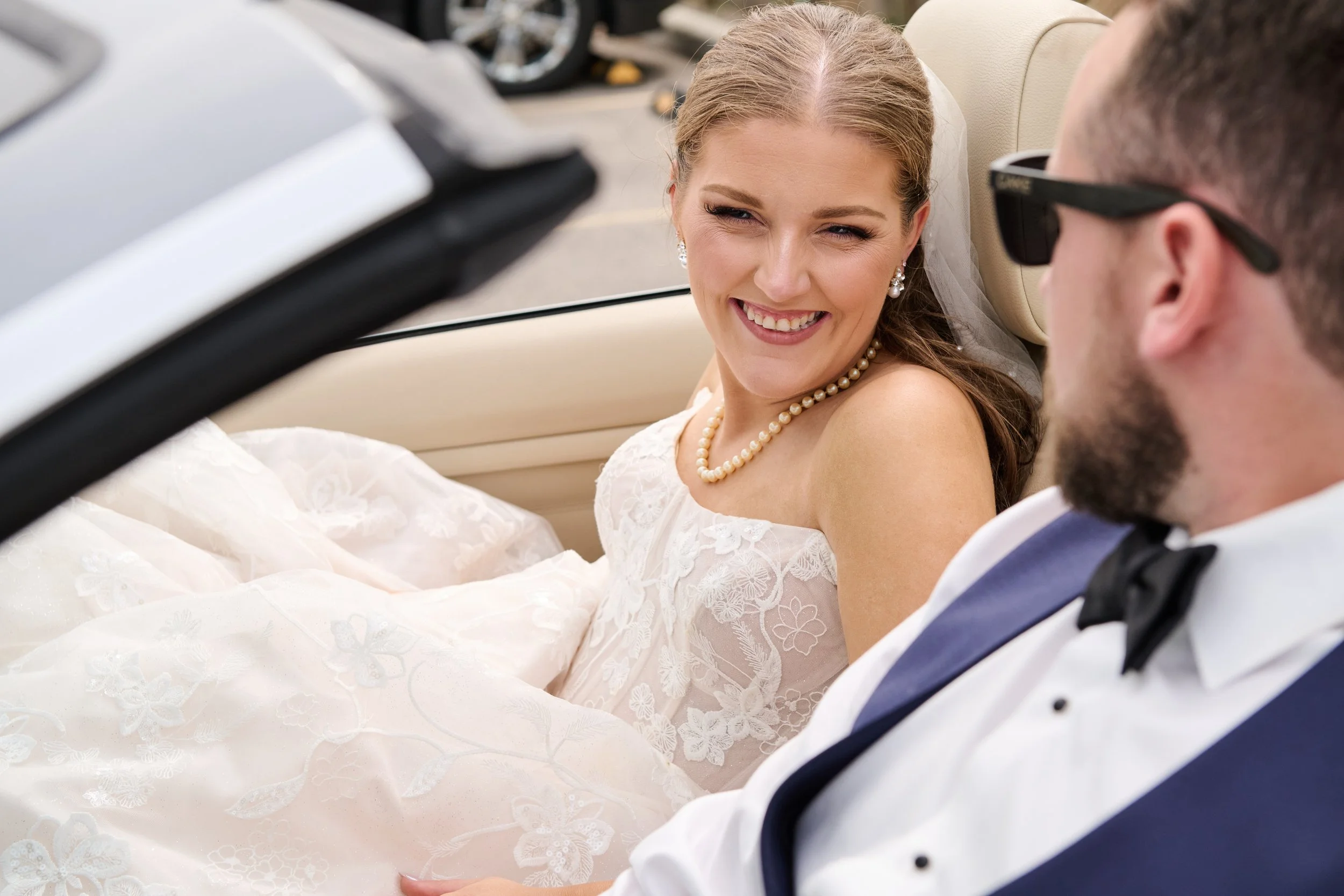 A bride and groom sitting in a convertible car, smiling and looking at each other.