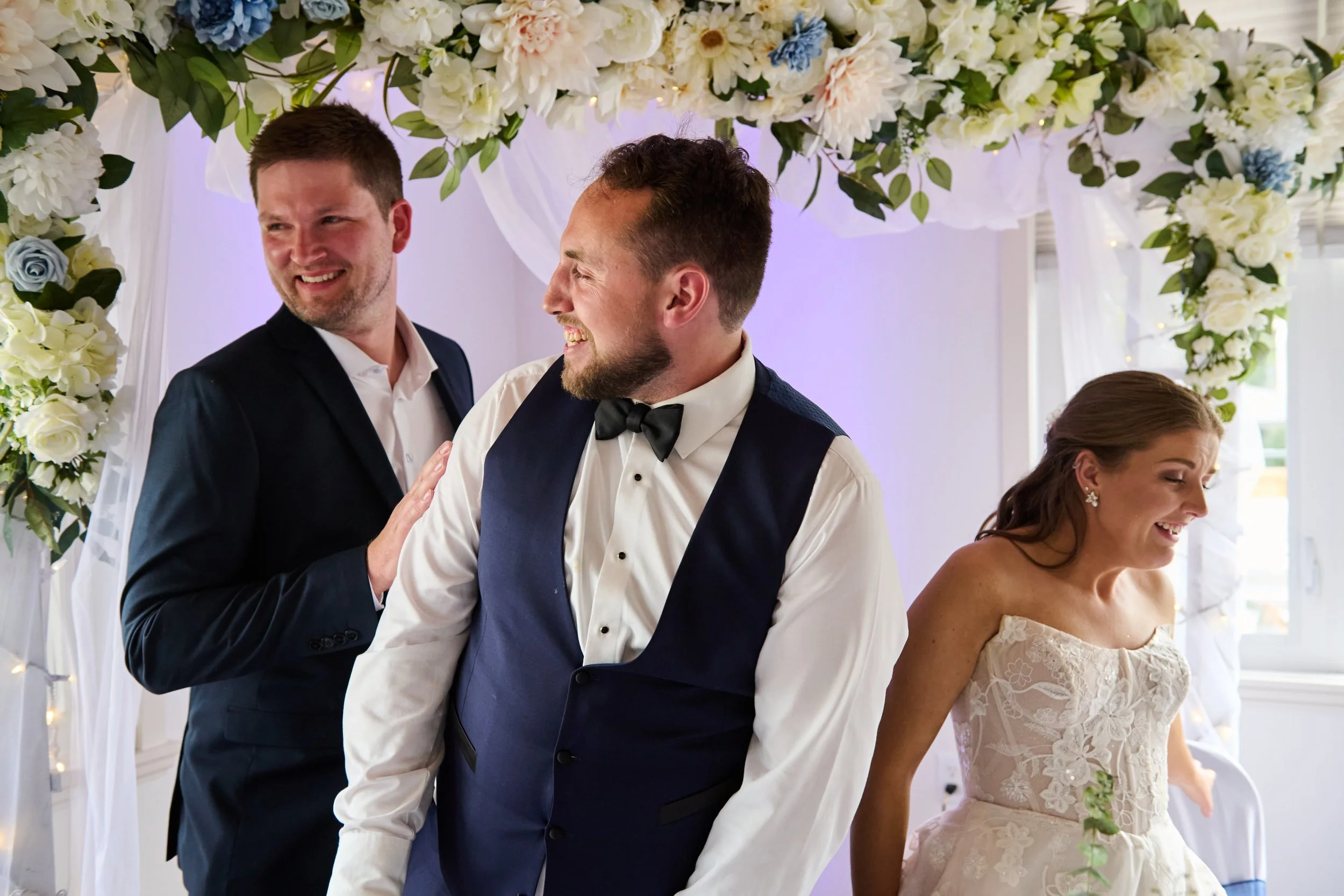 A wedding scene with four people, the bride and groom in the foreground and two men behind them, under a floral arch with white and blue flowers, white drapery, and fairy lights, smiling and laughing.