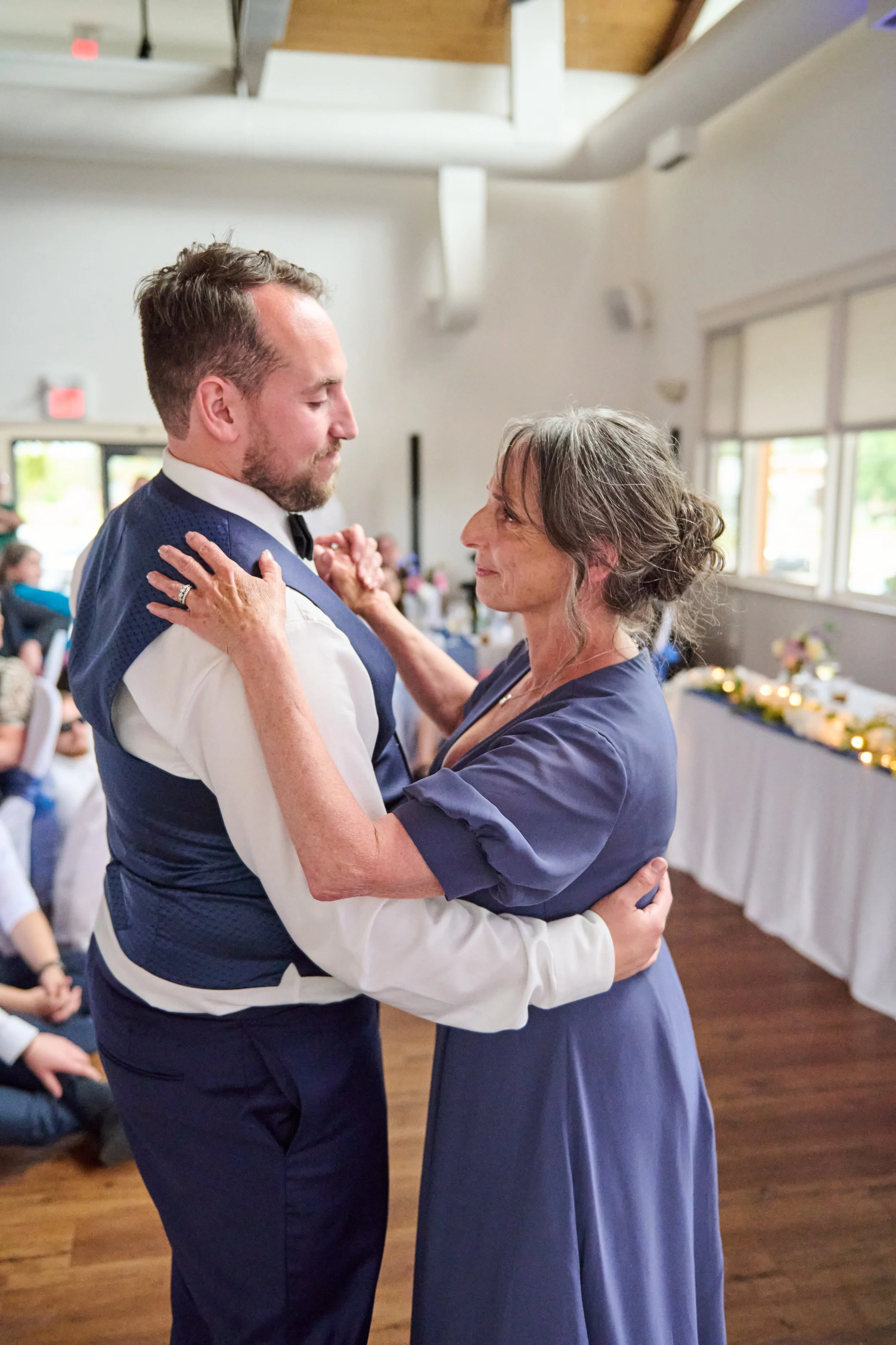A man in a tuxedo dancing with an older woman in a blue dress indoors at a celebration or wedding reception.