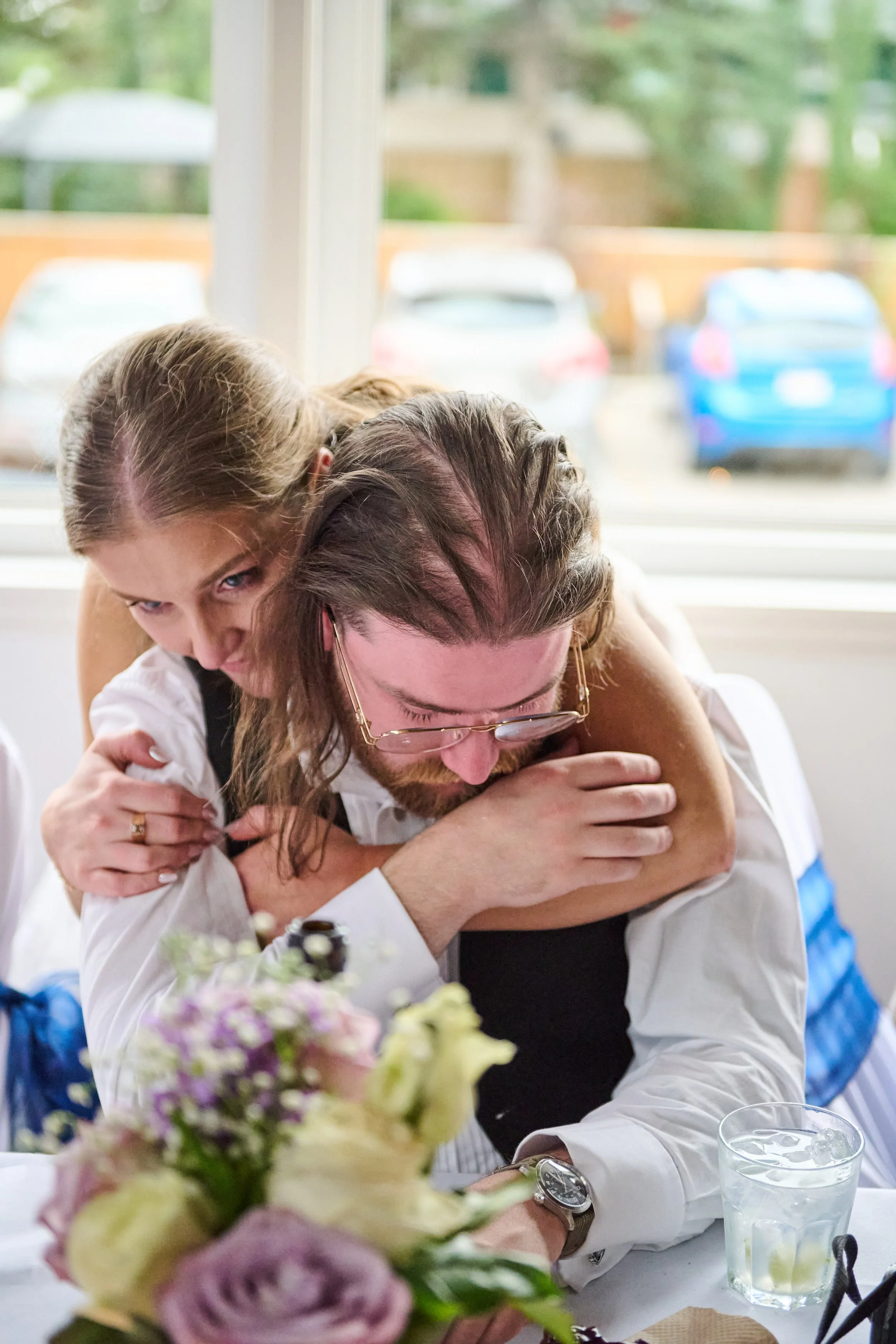 A woman and a man share a hug at a celebration, with a table of flowers and drinks in front of them and a window overlooking a parking lot outside.