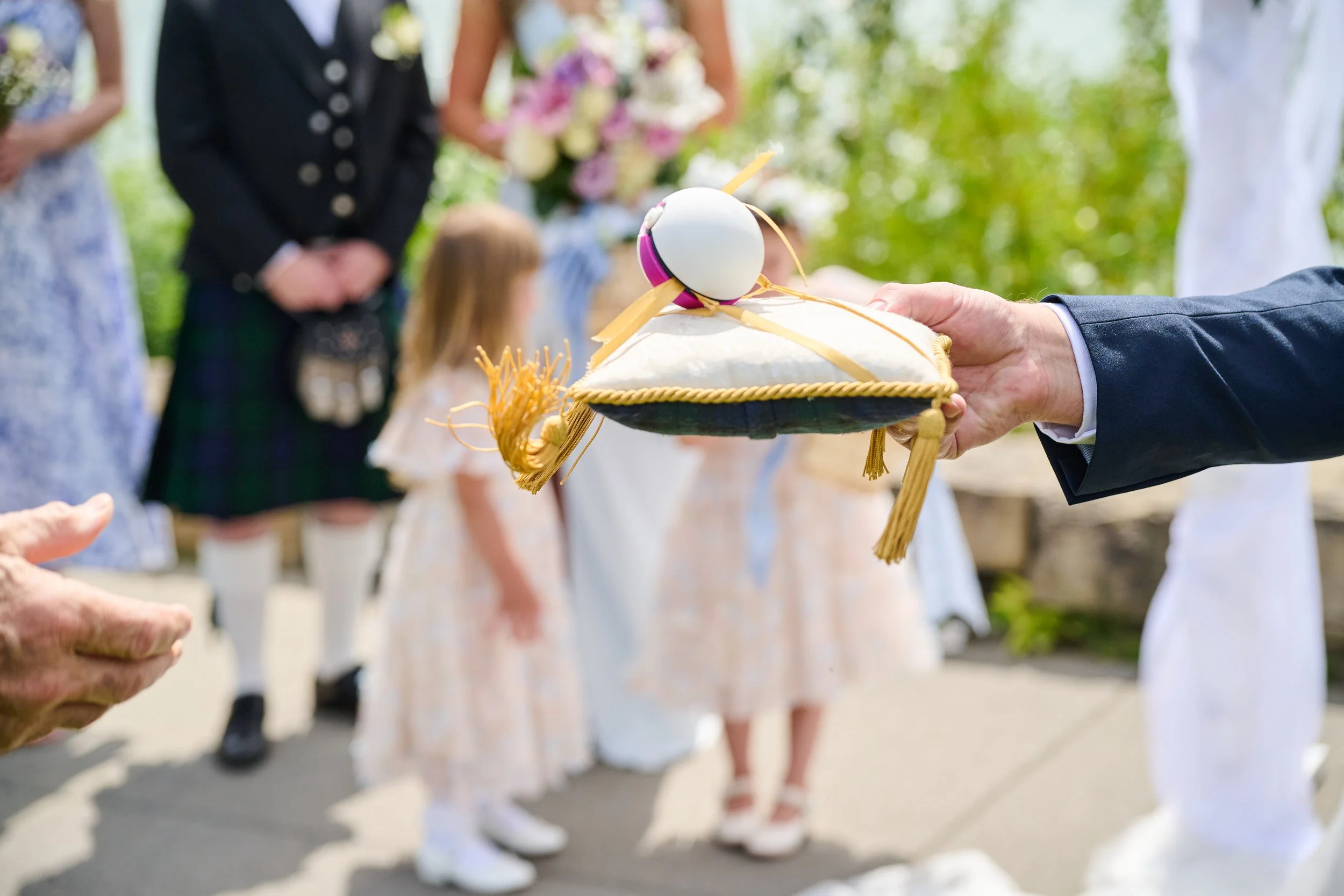 A person holding a decorated pillow with an egg and ribbon at a traditional outdoor wedding ceremony, with wedding guests and children in the background.