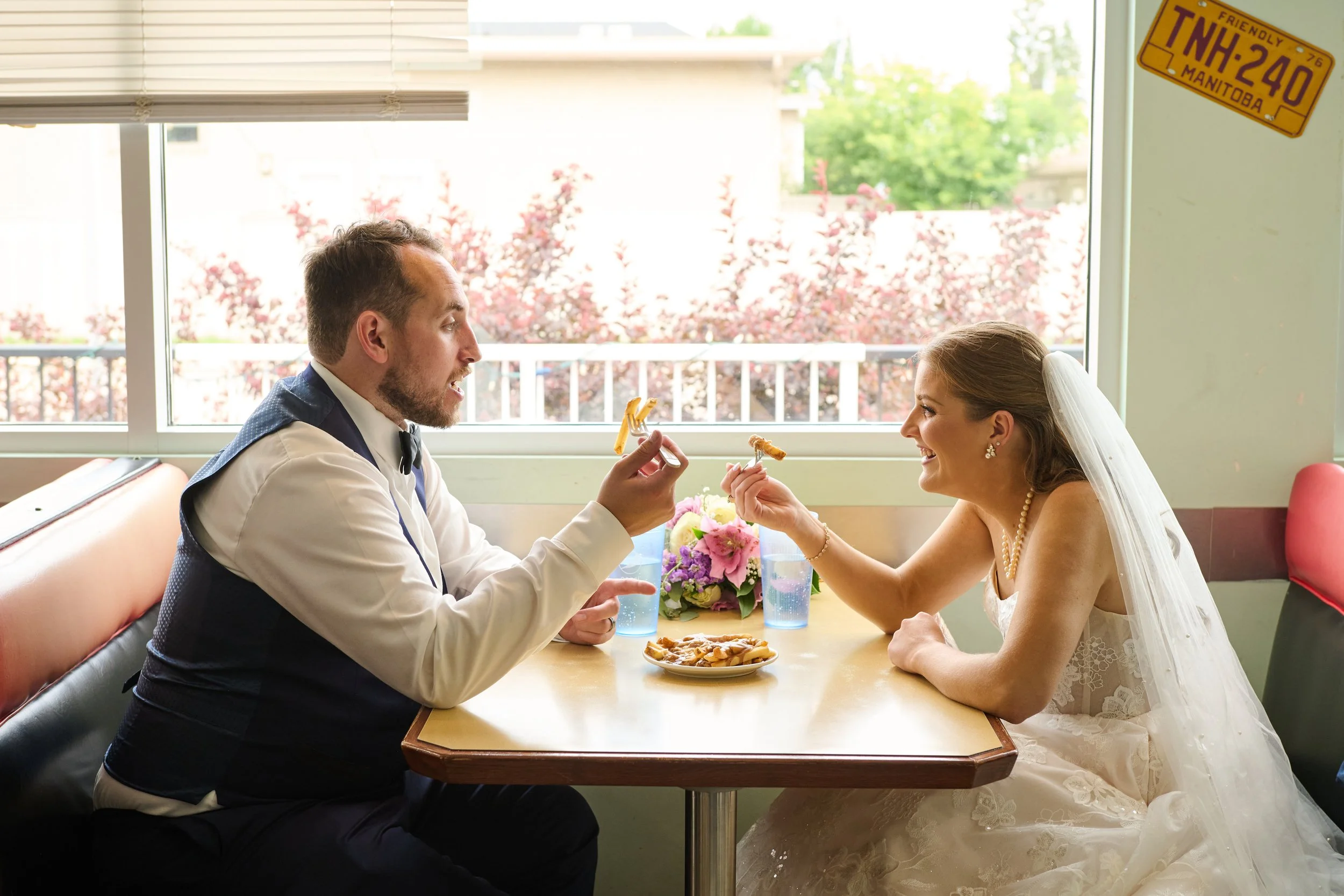 A newlywed couple in wedding attire sharing food at a restaurant table, with sunlight and pink flowering trees outside the window.
