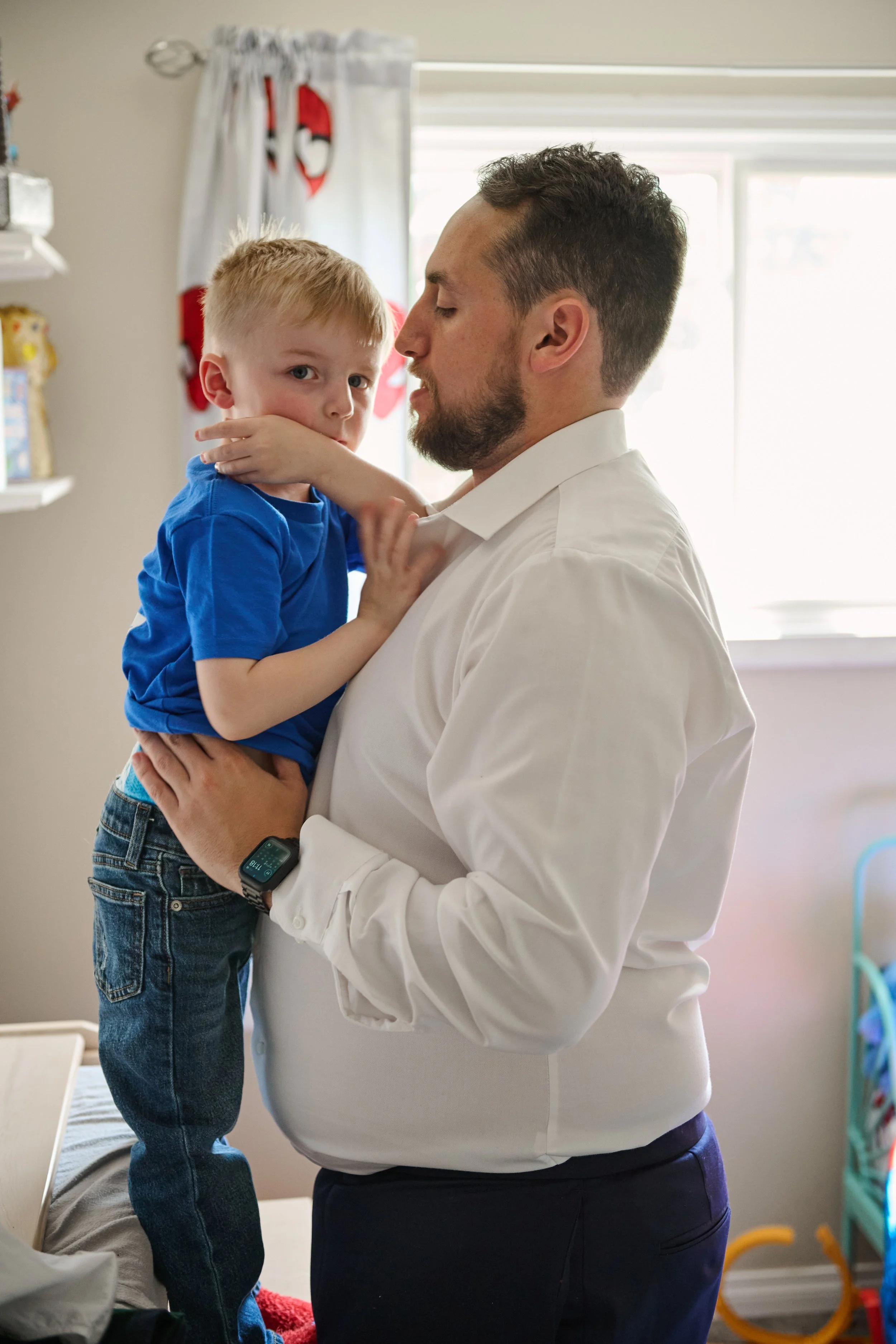 A man holding a young boy in a light-filled room. The boy has blond hair and is wearing a blue shirt and jeans. The man has dark hair and a beard, wearing a white shirt, a wristwatch, and dark pants. The room has a window with a white curtain decorat